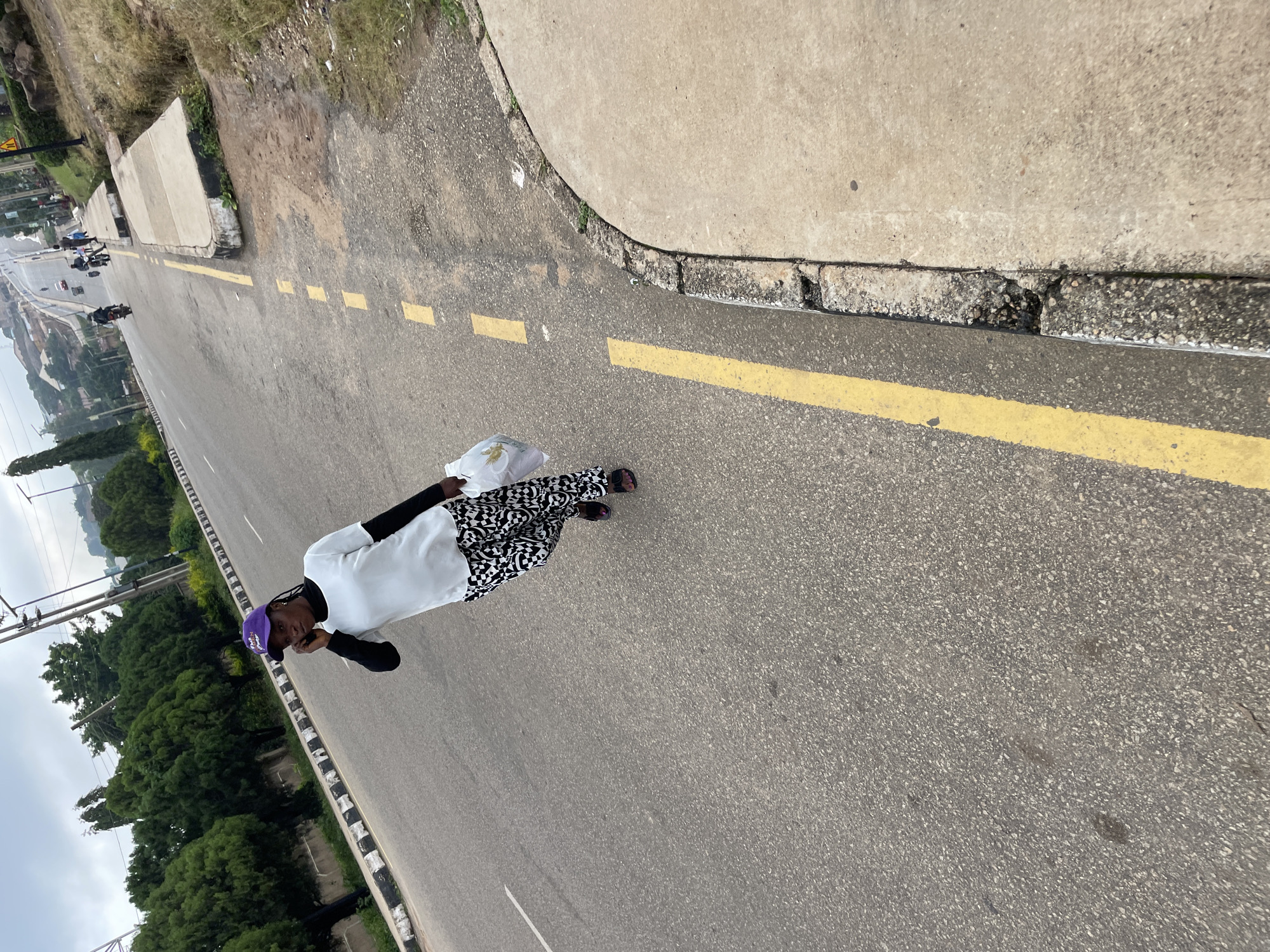 A woman in a white shirt and black and white patterned pants is walking on a road in Jos, Nigeria, while talking on a mobile phone. She is also carrying a white plastic bag in her right hand. The road has a curb and yellow painted lines. In the background, there are trees and buildings, and other people and vehicles can be seen on the road. The sky is overcast.