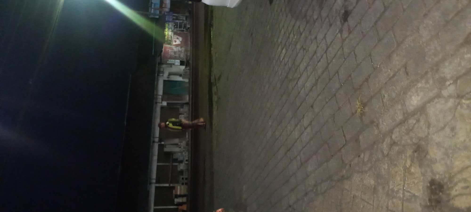 A person is walking on a paved area at night in front of a low-rise building that appears to be a row of shops. The person is wearing a light-colored reflective vest over a darker garment and pants. There is a bright light source to the right of the image, casting a beam downwards. The ground is paved with rectangular bricks. The building has a dark roof and lighter colored walls with open sections or doorways. There are some objects visible inside the building, possibly stacked items. The overall lighting suggests it is nighttime. The location is identified as Banjarsari, Indonesia.