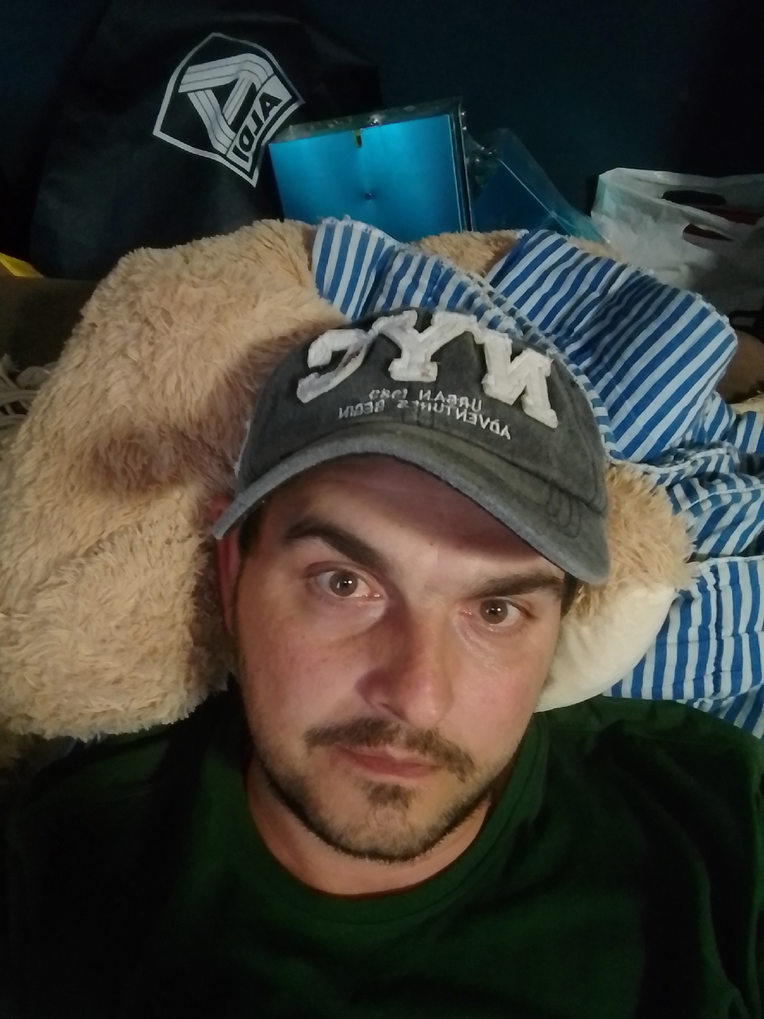 In a dimly lit indoor environment, a man wearing a dark green t-shirt and a grey baseball cap with "NY" embroidered on it is the primary subject. The cap also has text below "NY" that reads "URBAN ADVENTURES BEGIN". He has short brown hair, brown eyes, and a light beard and mustache. The man is positioned in the foreground, looking directly at the camera. Behind him, to his left, is a large, light brown, fluffy object that appears to be a stuffed animal. To the right of the stuffed animal and behind the man's head, a blue object with a metallic sheen is visible, partially obscured by what looks like a blue and white striped blanket or fabric. Further to the right, a white plastic bag is visible. The background is dark, suggesting the image was taken at night or in a poorly lit room. There is no visible indication of the specific location within the city of Wedel, Germany.