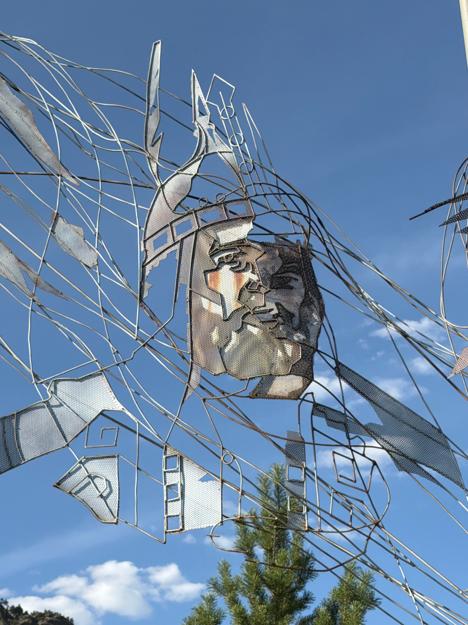 This is a close-up, upward-facing outdoor photograph of a metal sculpture against a bright blue sky with scattered white clouds. The sculpture is abstract and appears to be made of wire and perforated metal. A stylized human face is visible in the center of the frame, constructed from pieces of metal that suggest features like eyes, a nose, and a mouth. Surrounding the face is a chaotic arrangement of thin metal wires and geometric shapes formed from perforated metal, creating a sense of movement and fragmentation. In the lower part of the image, the tops of evergreen trees are visible, along with a hint of a rocky hillside in the far background. The metal of the sculpture shows some signs of rust and weathering. The location is identified as a rural district in Kazakhstan.