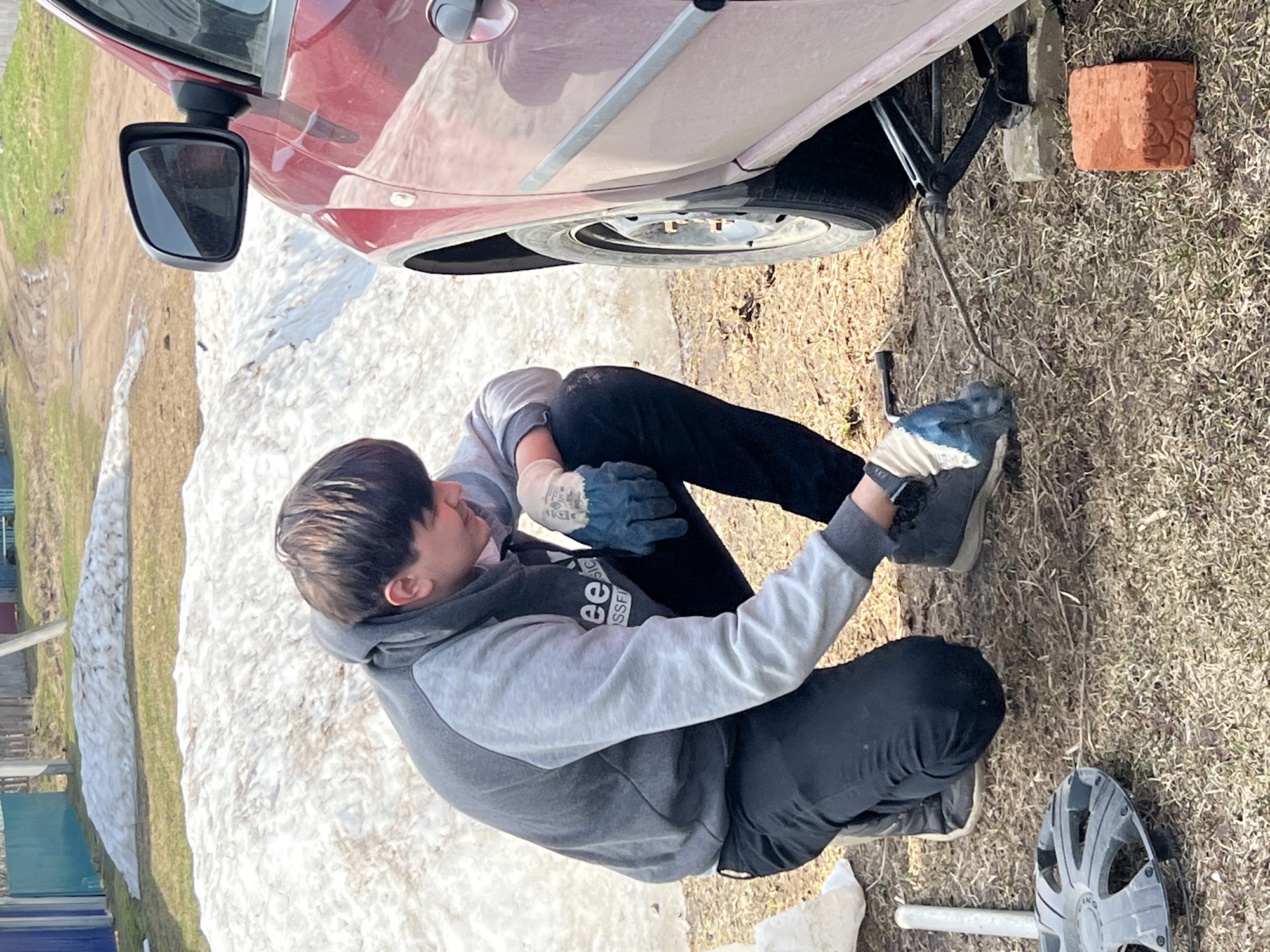 A young man is squatting next to a red car that is raised by a jack. The man is wearing a gray and black hoodie, black pants, gray shoes, and gloves. He appears to be operating the jack with a hand tool. The car's tire is visible, and a brick is positioned nearby. The setting is outdoors, with patches of snow, grass, and a house in the background. Based on available information, the location is Zhigalki, Russia.