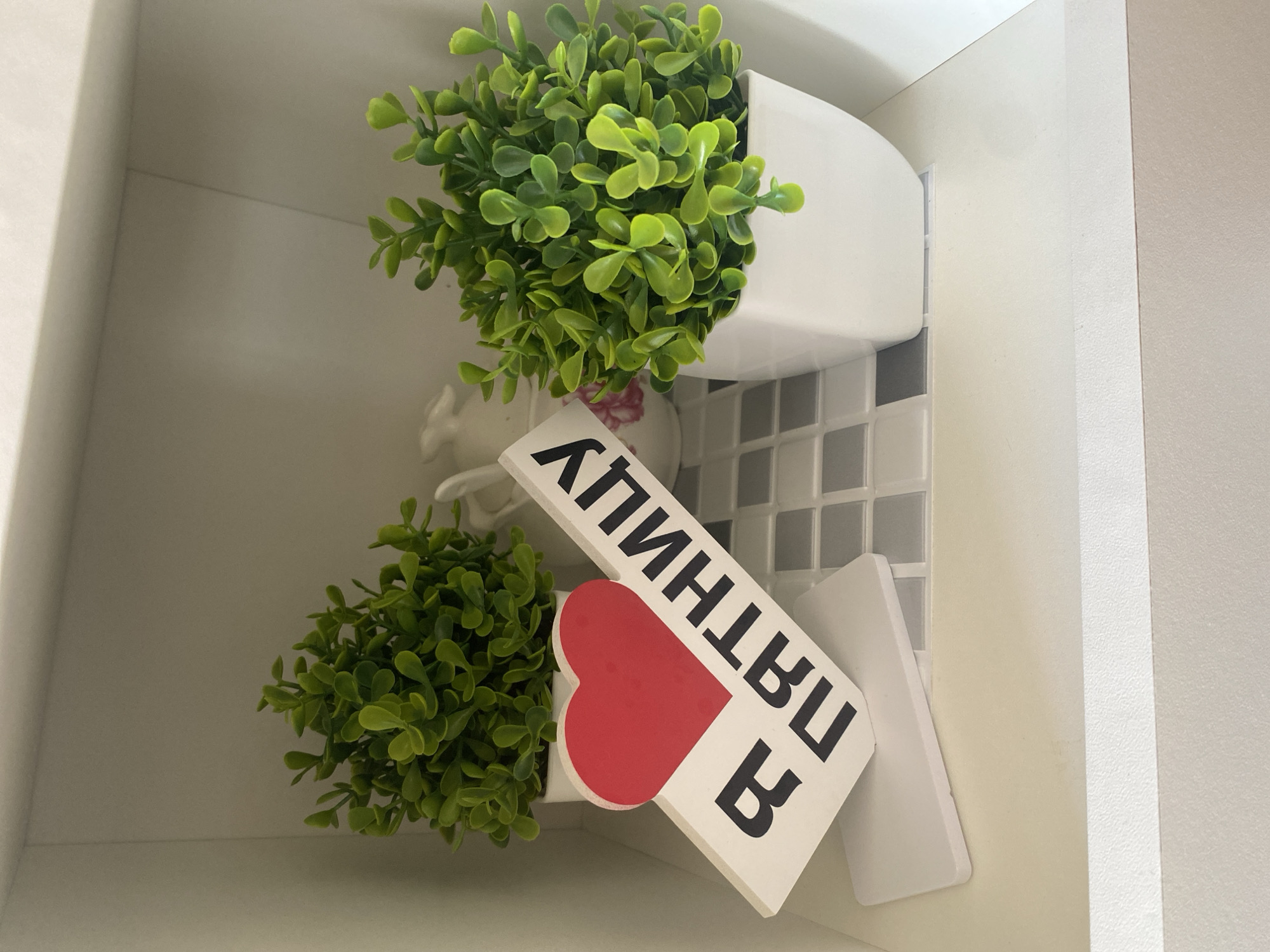 A close-up, eye-level shot shows a decorative sign with a red heart and Cyrillic text reading "Я ПЯТНИЦУ" on a white shelf. The sign is angled, with the heart at the top left and the text descending to the right. Behind the sign, on the same shelf, are two artificial green plants in white square pots. To the left, a smaller plant sits behind the sign. To the right, a larger, fuller plant occupies its white pot. A patterned tiled surface, featuring alternating grey and white squares, is visible behind the sign and between the plants. Further back and slightly out of focus, a white ceramic object, possibly a pitcher or vase with a floral design, is present. The shelf is part of a white shelving unit. The overall lighting is bright and even. The image does not depict the city of Chaikovsky, Russia.