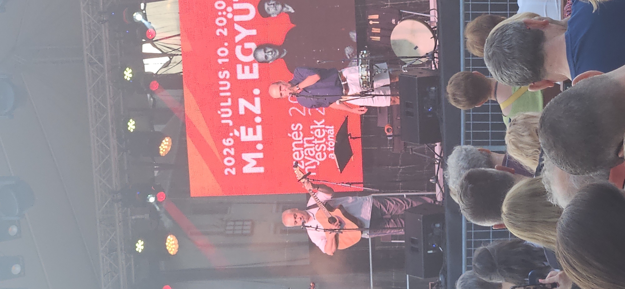The image shows a live music performance on an outdoor stage in Hévíz, Hungary. A band, "M.É.Z. Együttes", is performing in front of a large red LED screen displaying text that includes "2026. JÚLIUS 10. 20:00". The band members are visible on stage. One man is playing an acoustic guitar and singing into a microphone. Another man is singing into a microphone and wearing a blue shirt and white shorts. Two other band members are visible in the background, wearing dark shirts. The stage is equipped with speakers and lighting equipment. A crowd of people is visible in the foreground, watching the performance. The event appears to be taking place during the evening, given the stage lighting.