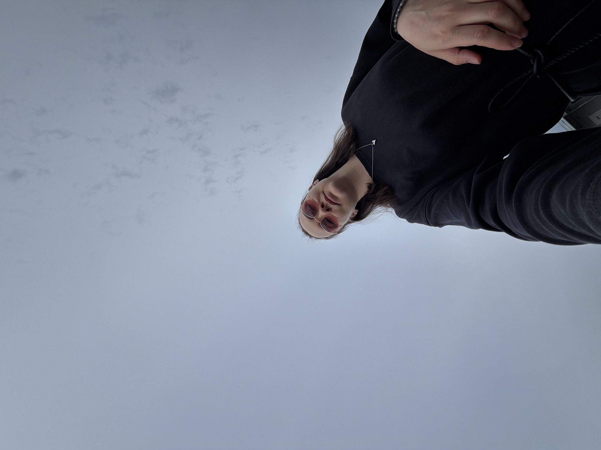 A low-angle, tilted photograph shows a person with fair skin, wearing round, rose-tinted glasses and a black outfit, including what appears to be a sweater and possibly a jacket. The person has long dark hair and is wearing a silver necklace. The background is a grey sky with faint clouds visible. The location appears to be outdoors, but no specific architectural or geographical features are discernible to place it in Chaikovsky, Russia, or any other specific location. There are no discernible actions or interactions taking place within the photograph.