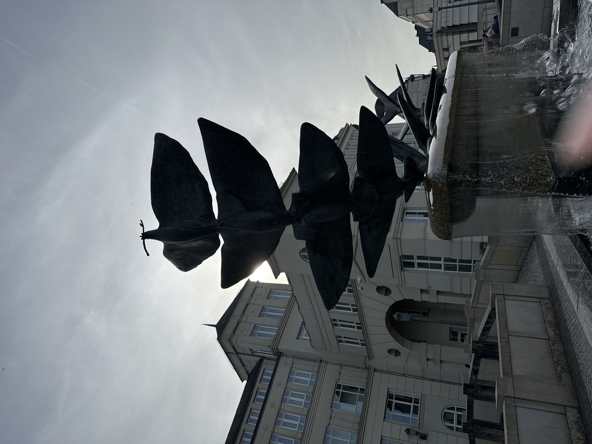 A dark, abstract sculpture of five bird-like figures in flight, stacked vertically, dominates the foreground of the image, silhouetted against a bright, overcast sky. The sculpture is part of a fountain, with water cascading down its sides and splashing into a pool below. Behind the sculpture, a light-colored, multi-story building with numerous windows and an arched entrance is visible. The building appears to be modern architecture. To the right of the building, another building with a pitched roof and dormer windows can be seen. Paved walkways and low concrete barriers form a plaza around the fountain. The ground is wet from the fountain's spray and scattered with what appears to be frost or ice, suggesting cold weather. In the far right of the image, two people are seated on a ledge. The overall scene suggests an outdoor public space in a city, likely Luxembourg City, Luxembourg, given the architectural style and the presence of a water feature.