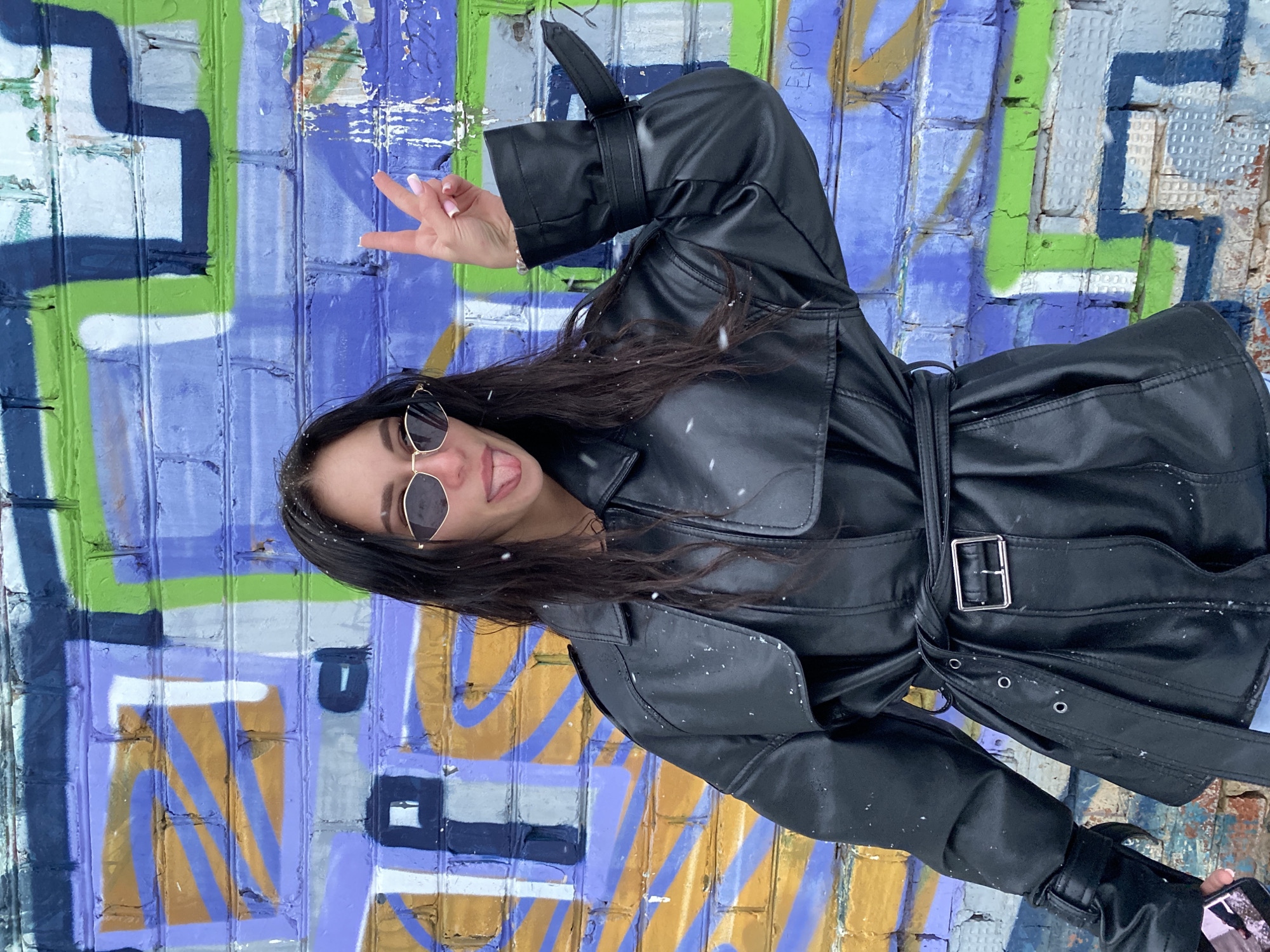 A woman with long, dark hair wearing sunglasses and a black leather trench coat stands in front of a brick wall covered in graffiti. She holds up her hand with a peace sign and sticks out her tongue. Light snowfall is visible in the air and on her coat. The graffiti on the brick wall features a mix of colors including green, blue, purple, yellow, and white. The graffiti style includes geometric shapes and abstract designs. The picture was taken in Chaikovsky, Russia.