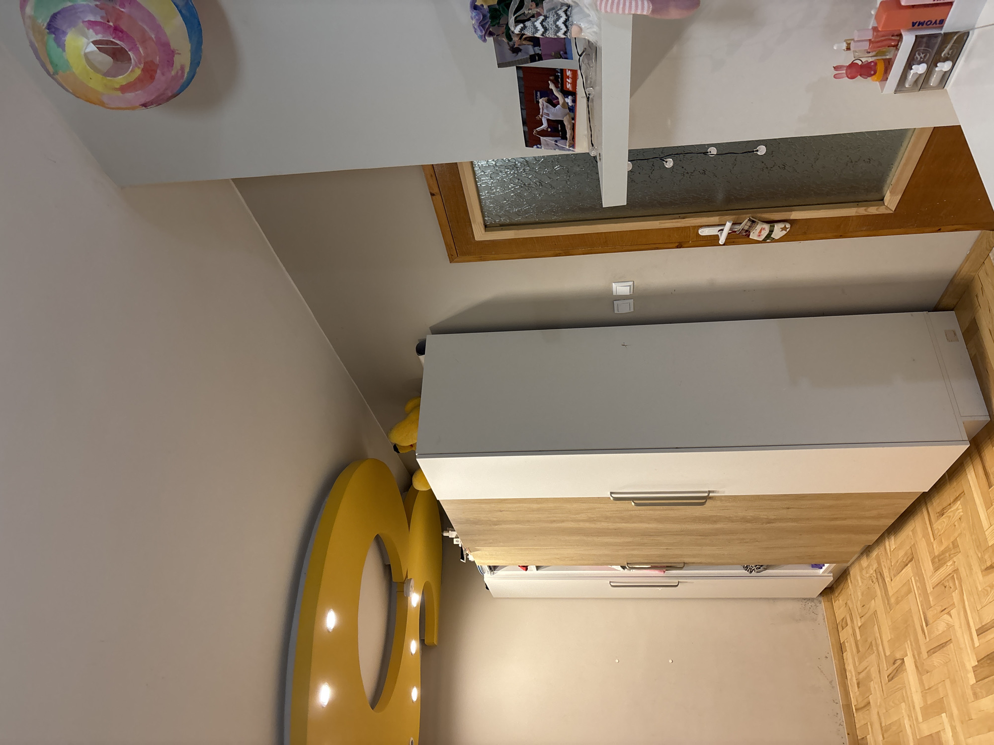 A rotated interior shot shows a bedroom corner with a light grey wall, a yellow letter "G" shaped light fixture, and a dresser with white and wood-grain drawers. In the upper part of the image is a dormer window, with an attached shelf featuring decorative items and a window with a wood frame. A circular, multicolored wall decoration is attached to the ceiling. The location is possibly in a residential building in Sofia, Bulgaria, based on the common European architectural style and the presence of dormer windows.