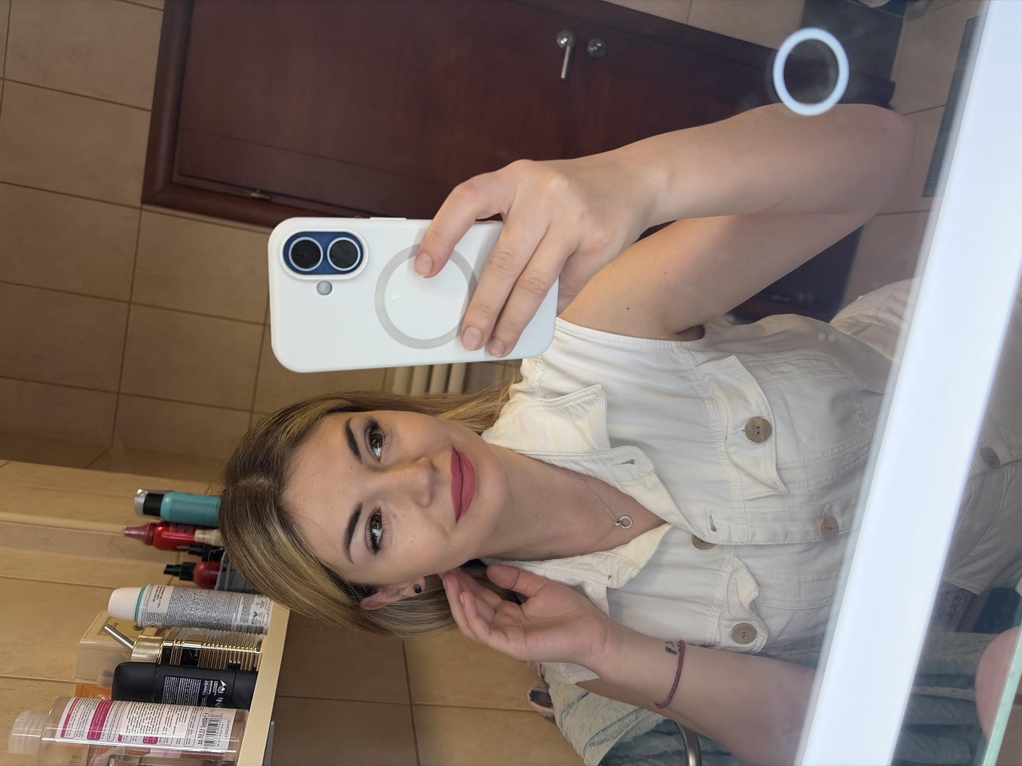 A woman is taking a selfie in a bathroom. She holds a white phone in her right hand. The bathroom has beige tile walls and a dark wood door with a silver handle. A shelf on the left holds toiletries and hair products. The woman wears a white button-down shirt and a silver necklace. She is looking into the camera with a slight smile. The photo appears to be taken in a mirror with a ring light reflection in the top right corner. No information in the image specifically indicates the location is Budva, Montenegro.