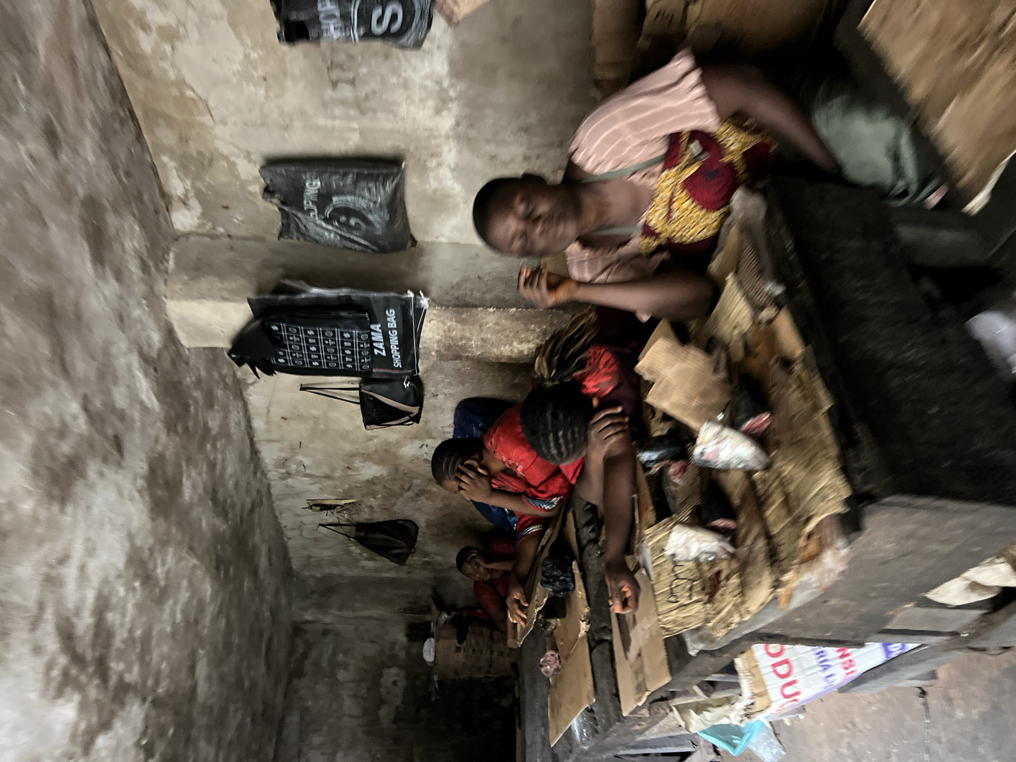 In Nkpor, Nigeria, four women are seated at a rough wooden stall in a dimly lit indoor market. The walls and ceiling are made of rough concrete with visible signs of wear and possibly mold. The women appear to be vendors, with some resting their heads on their hands, suggesting fatigue or boredom. On the stall in front of them, there are various items, including what appear to be dried fish, some wrapped in plastic, and other unidentifiable objects. Bags are hanging on the wall behind them, some with writing that suggests they are shopping bags. The overall atmosphere is one of a humble, everyday marketplace.