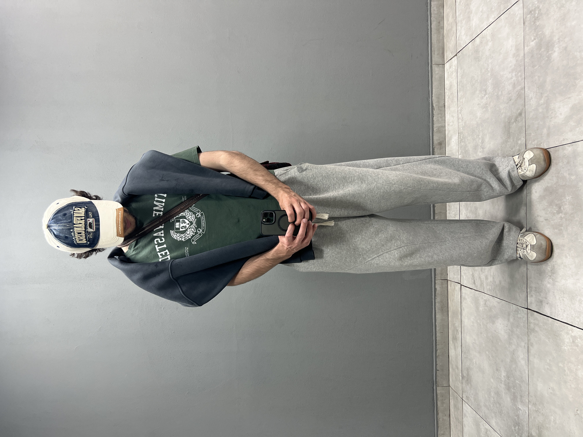 A person stands against a gray wall and tiled floor. They wear a baseball cap, a green t-shirt, gray sweatpants, and sneakers. A dark gray jacket is draped over their shoulders. They hold a smartphone in their hands. The location is likely an indoor setting in Yerevan, Armenia.