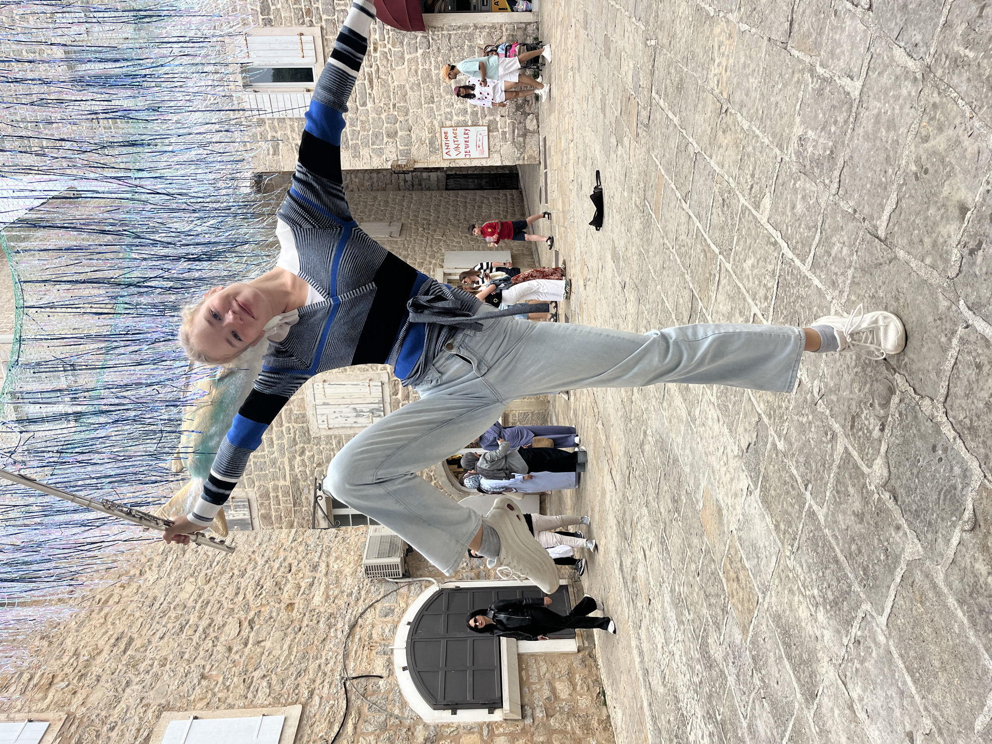 A woman stands in a dynamic pose in what appears to be a cobblestone square in Budva, Montenegro. She balances on one leg, extended to the right, and holds a flute in her left hand. Her right arm extends upward. She is wearing a striped long-sleeved shirt, light-colored jeans, and white sneakers. Her hair is blond.
The background consists of old stone buildings with closed windows and doorways. A group of people is visible in the distance, seemingly tourists. A storefront with a sign that reads "ANTIQUE VINTAGE JEWELRY" is visible. In the foreground, a woman in a black leather jacket stands near a doorway. Overhead, a shimmering decoration with metallic streamers hangs. The cobblestone pavement suggests an old city square.