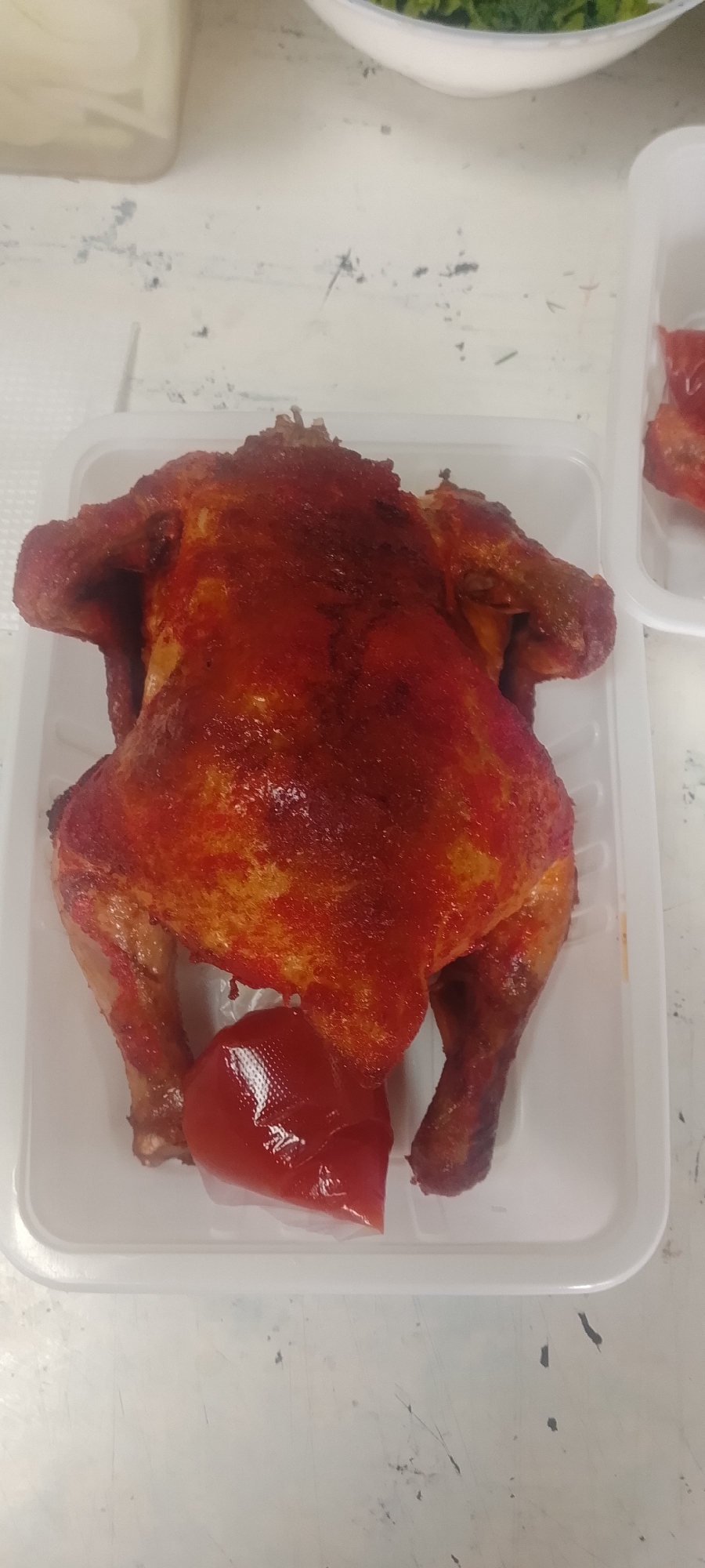 A whole roasted chicken sits in a white plastic takeout container, its skin a rich, reddish-brown. A small packet of red sauce, likely ketchup, is placed in front of the chicken. The container is on a light-colored, speckled surface that appears to be a table or countertop. The background is blurred, but suggests a domestic or commercial kitchen environment. The scene is lit from above. There are no people visible in the image. This image was taken in Aydarken, Kyrgyzstan.