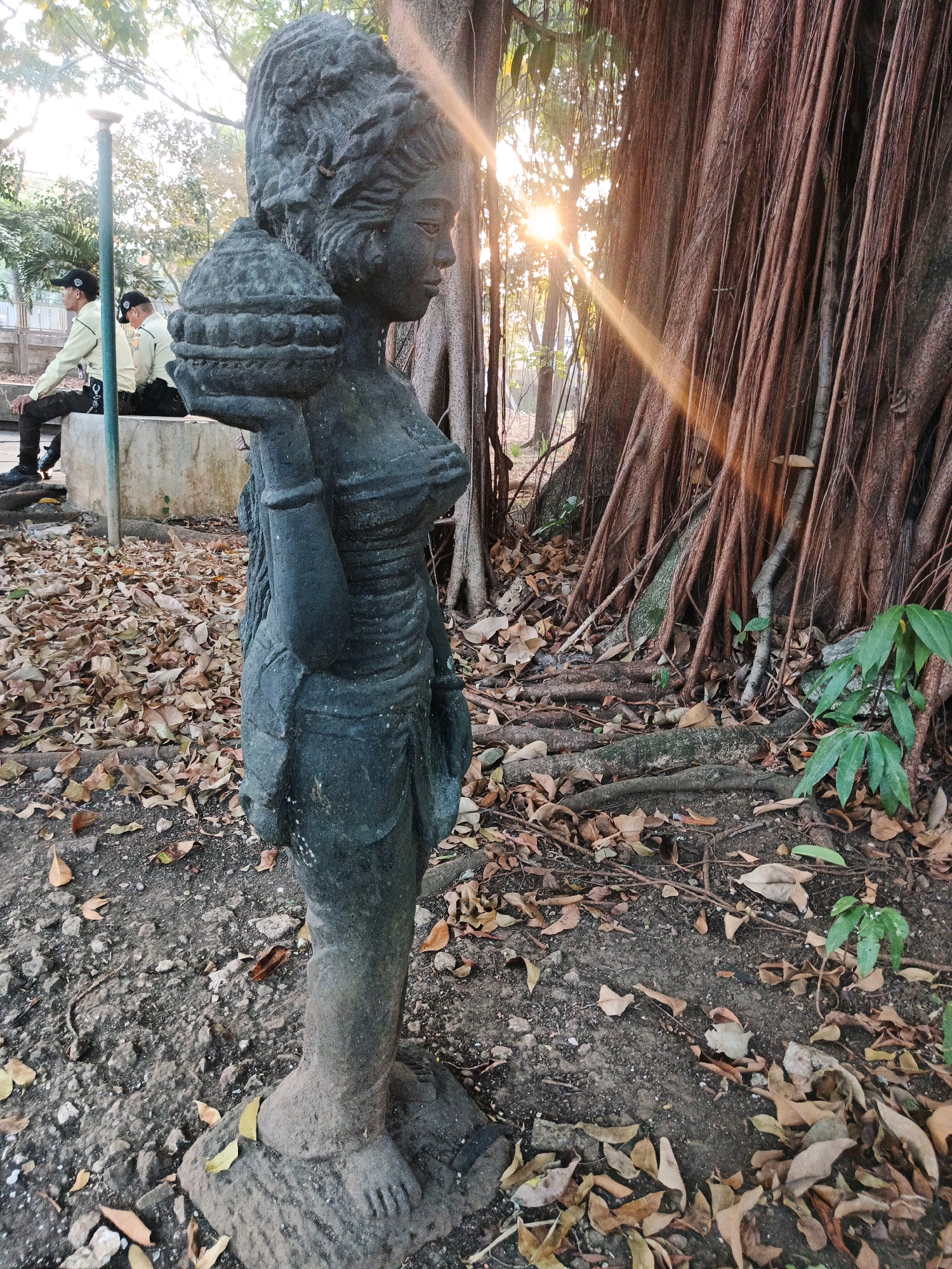 The image depicts a weathered stone statue of a woman in the foreground, positioned outdoors on a ground covered with fallen leaves and small stones. The statue is dark in color and appears to be ancient, with intricate details in its headdress, clothing, and the object it carries on its shoulder. Behind the statue, a large, old banyan tree with aerial roots is prominent. Sunlight filters through the leaves of the trees in the background, creating a lens flare effect and casting beams of light. In the middle ground, two individuals in uniform, possibly security guards, are seated on a concrete bench to the left. They are wearing light-colored long-sleeved shirts, dark pants, and caps. A lamp post is also visible near the bench. The overall environment suggests a park or a historical site. This scene is located in the Bandung City, Indonesia, specifically within the grounds of the Geological Museum of Bandung.