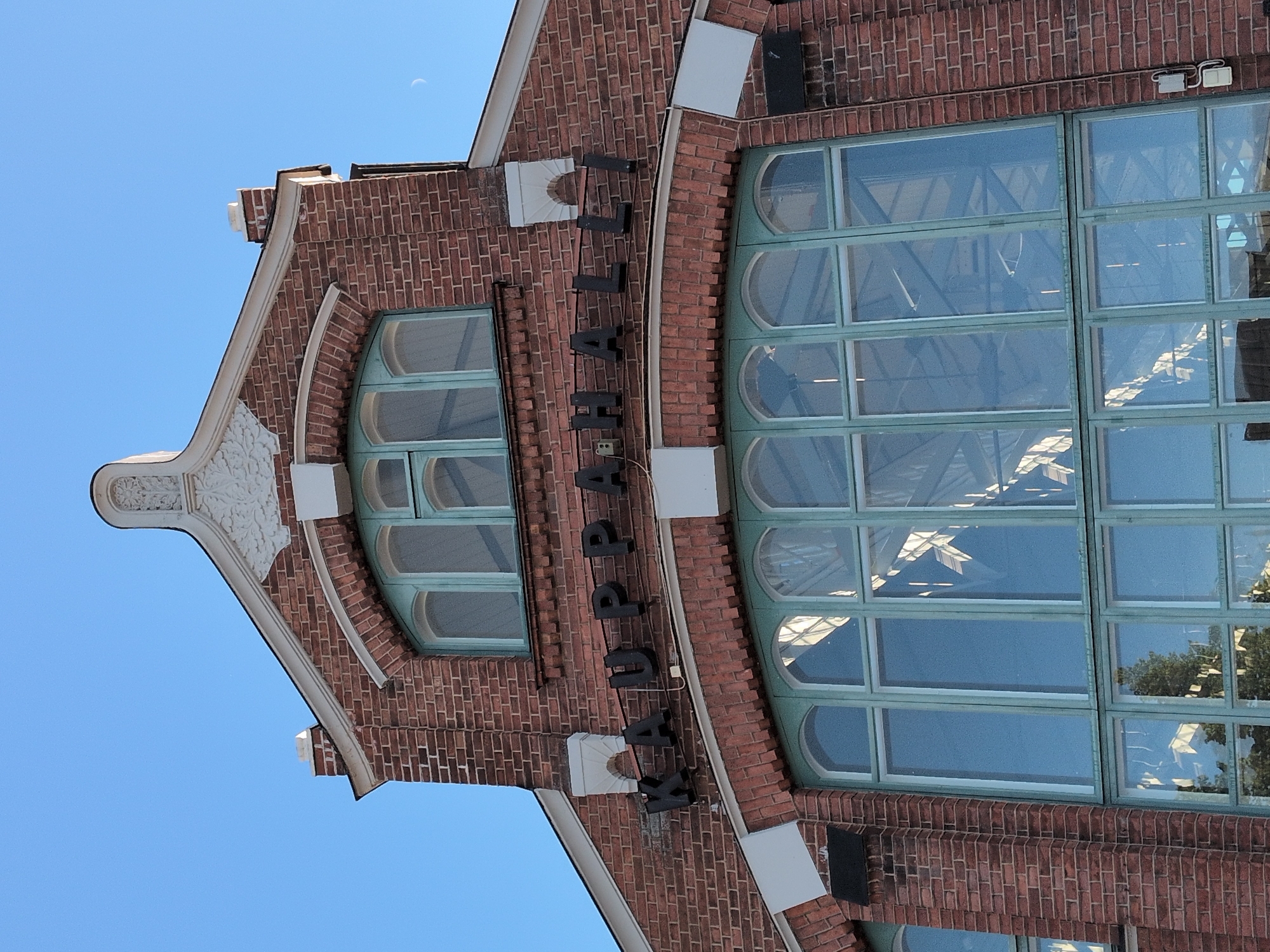 The image shows the facade of the Oulu Market Hall (Oulun Kauppahalli), a historic building located in the city of Oulu, Finland. The facade is constructed of red brick and features large, arched windows with green frames. Above the windows, the word "KAUPPAHALLI" is visible in black, three-dimensional lettering. The sky is clear and blue, with a crescent moon visible in the upper right quadrant of the image. Architectural details, such as decorative white elements above the windows and a chimney, are also present. The overall impression is of a well-maintained, classic building against a bright sky.