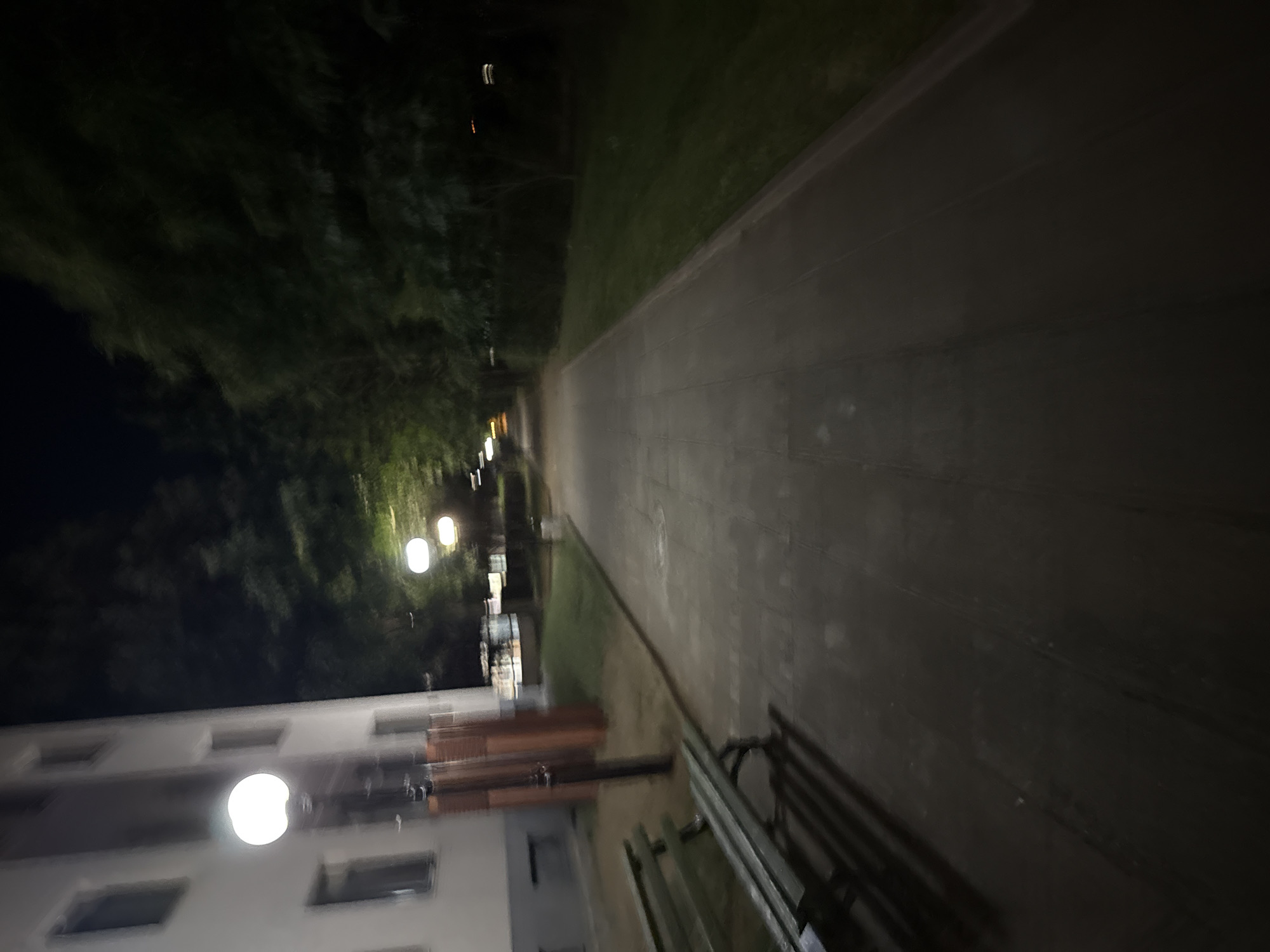 The image depicts an outdoor scene at night in Blagoevgrad, Bulgaria. It appears to be a sidewalk or paved pathway lined with trees and grass. A building is visible on the left side of the image, with windows and a light fixture. A bench is situated on the right side of the image along the pathway. The lighting is dim, with streetlights illuminating the path and building. The overall impression is a quiet, possibly residential area at night.
