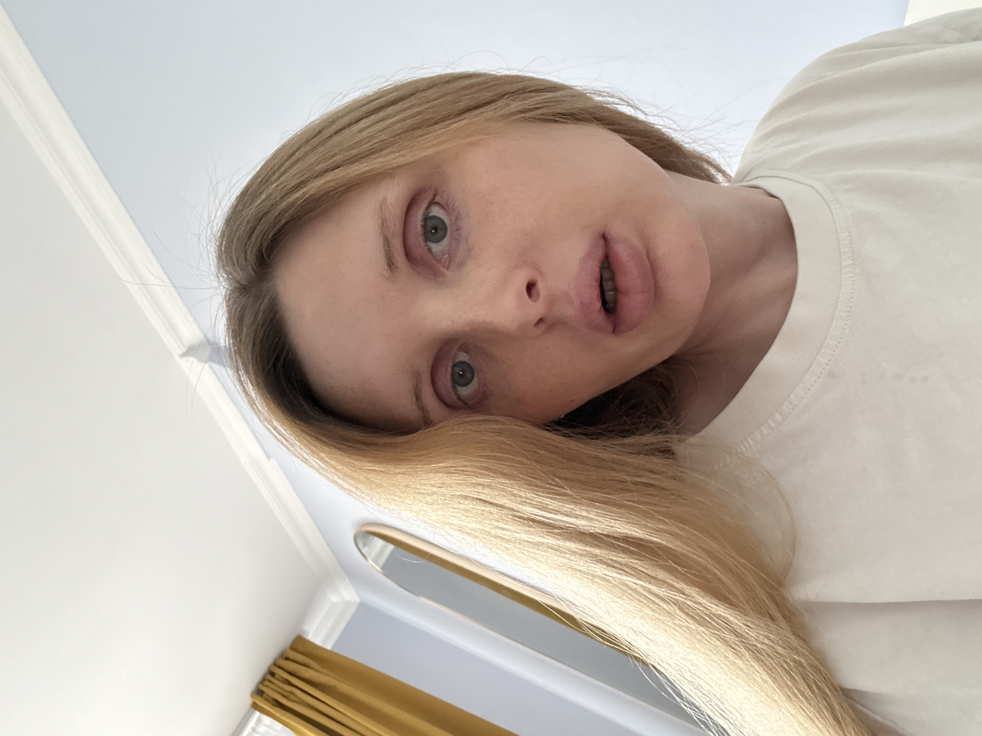 A young blonde woman is positioned in the foreground, her face angled towards the camera. She is wearing a light-colored t-shirt. Her eyes are open, and her mouth is slightly ajar, revealing braces on her teeth. Her hair is long and blonde, falling around her face and shoulders. Behind her, the background consists of a light blue wall with white crown molding. To the left, a yellow curtain is visible, and a tall, oval mirror with a gold frame hangs on the wall. The ceiling is white. The image appears to be a selfie, taken indoors.