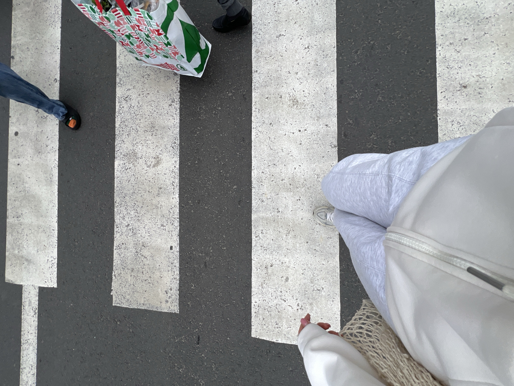An overhead shot captures a pedestrian crossing in Ternopil, Ukraine, with multiple people traversing or standing on the black asphalt marked with thick white stripes. In the foreground, the viewer's legs are visible, wearing light grey sweatpants and white sneakers, along with a white jacket and a beige mesh bag. A hand, possibly the viewer's, shows light pink nail polish. To the left, another person wears blue jeans and dark shoes with an orange detail. Further into the frame, a person carries a white shopping bag adorned with a festive green and red pattern, which also displays red text that looks like "购" and a "G" in a circle. Another person's dark shoes and pants are visible near this shopping bag. The scene depicts an everyday activity on a street under what appears to be bright daytime conditions.