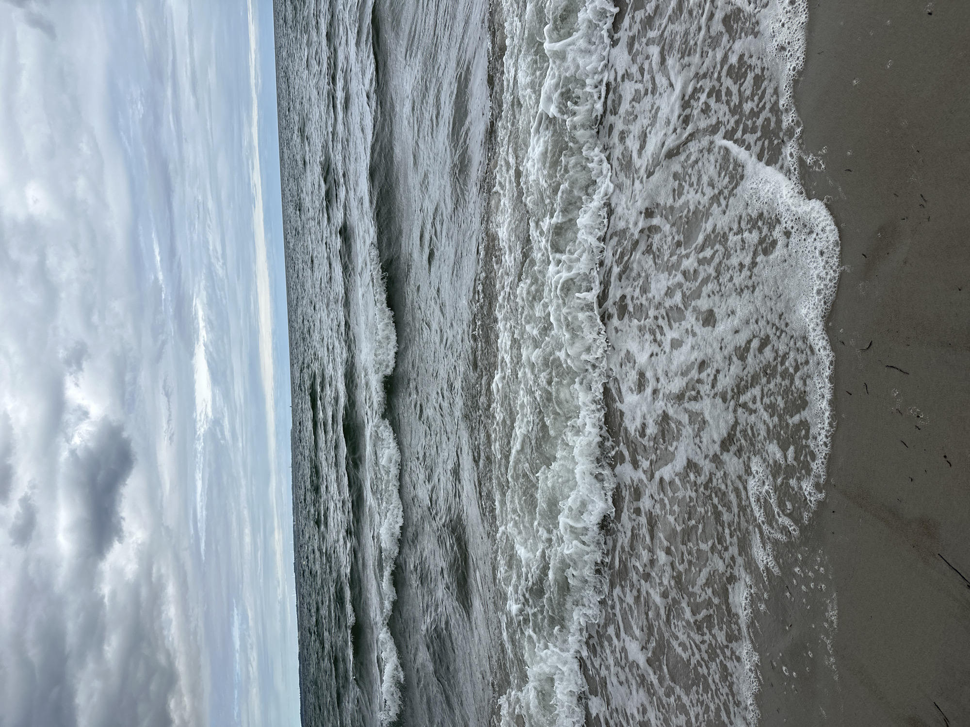 The image shows a view of the Baltic Sea from the beach in Scharbeutz, Germany. The sky is overcast with gray clouds. The sea is choppy with waves breaking on the shore, creating white foam. The beach is made of sand and there are some scattered pieces of seaweed. There are no people or objects visible in the image, except for the natural elements.