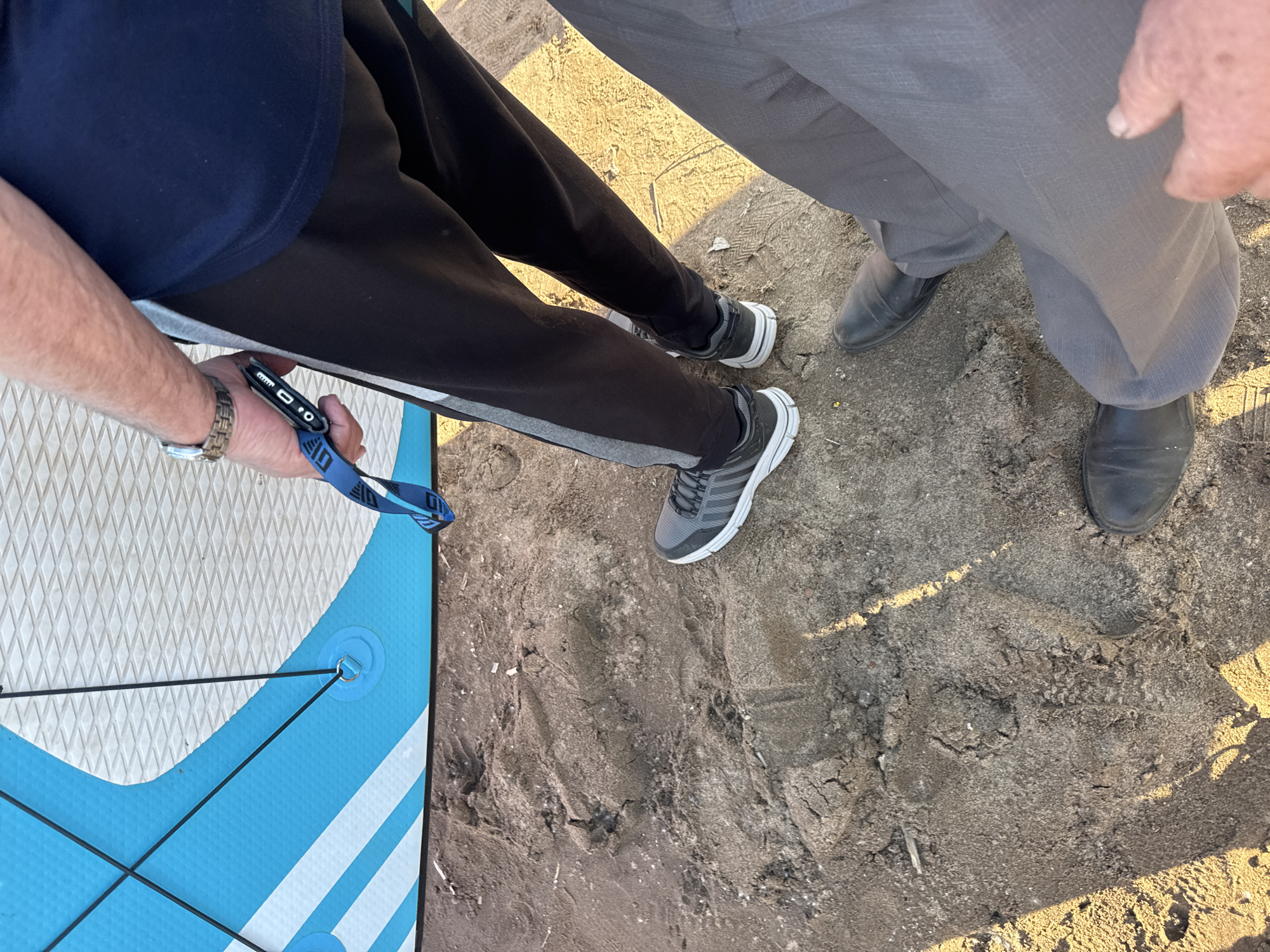This is a close-up, overhead shot focusing on the lower legs and feet of at least two individuals standing on a sandy surface, with a blue and white paddleboard partially visible on the left. The scene appears to be outdoors, with the lighting suggesting it is either early morning or late afternoon due to the long shadows cast on the sand. The sand is a dark, wet color and shows some texture and small debris, with visible footprints and disturbances.
One person is wearing black sweatpants with a gray stripe down the side and dark athletic shoes with white soles. They are holding a blue lanyard with a small black device attached, possibly a key fob or a tool. A watch with a metallic band is visible on their wrist.
Another individual, or possibly the same person from a different angle, is wearing gray dress pants and black dress shoes. Their attire suggests a contrast in activities or purpose compared to the person in athletic wear.
The paddleboard, positioned on the left side of the frame, is bright blue and white with a textured grip pattern. Black bungee cords are attached to a metal ring on the paddleboard, indicating it's prepared for use or storage.
The overall impression is one of preparation for or transition from an outdoor activity, possibly involving the paddleboard, on a sandy shore or beach. The lack of distinct facial features or interactions between the individuals makes it difficult to infer emotions beyond a sense of grounding and perhaps the anticipation of an activity. The setting is likely a location with access to water given the paddleboard.