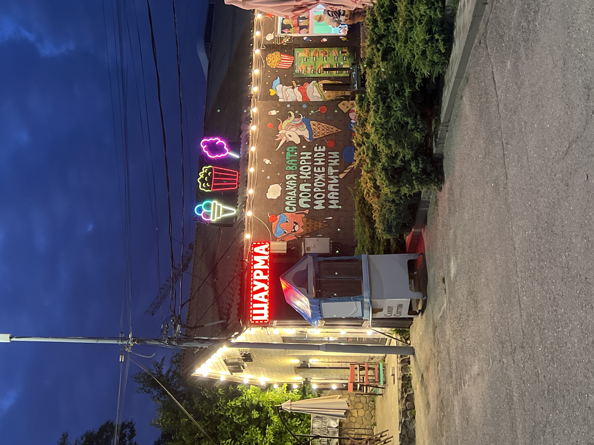 This is a medium-shot, eye-level, outdoor photograph taken at dusk of a food establishment in Krasnodar, Russia. The building, which appears to be a small storefront or kiosk, is decorated with bright lights and neon signs. A prominent red neon sign above the entrance reads "ШАУРМА" (Shaurma), indicating a food vendor specializing in this dish. To the left of the entrance, string lights illuminate the facade. In front of the building, there is a light blue and white kiosk with a sign that reads "Candy Cotton".
The wall of the building is covered in murals depicting various treats and cartoon characters. Visible images include neon outlines of an ice cream cone, a popcorn bucket, and cotton candy. Further down the wall, a mural features a unicorn in an ice cream cone, a stack of ice cream cones, and a popcorn bucket. A menu with prices in rubles is posted to the right of the murals.
The scene is surrounded by greenery, with dense bushes in the foreground and trees to the left. The ground is paved with asphalt, and there are some outdoor seating areas visible to the left, with umbrellas and chairs. The sky is dark blue, suggesting it is twilight. A tall utility pole with numerous wires is visible to the left of the building. A person can be seen through a window on the right side of the building, potentially an employee. The overall atmosphere is festive and inviting, typical of a casual dining or snack establishment in a recreational area.