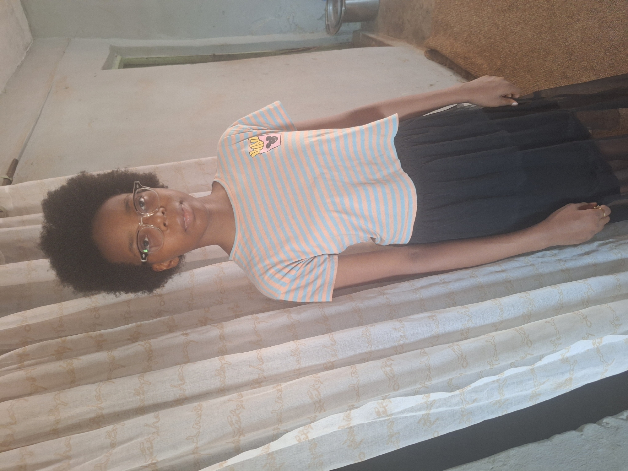 A young Black woman with an afro and glasses lies on a patterned off-white surface, possibly a bed or a draped area. She wears a striped peach and light blue t-shirt and a black skirt. The background includes a light-colored wall and a glimpse of a brown or tan surface in the upper right corner, and some piping can be seen by the wall. It cannot be ascertained from the image that the location is Sabon Tasha, Nigeria.