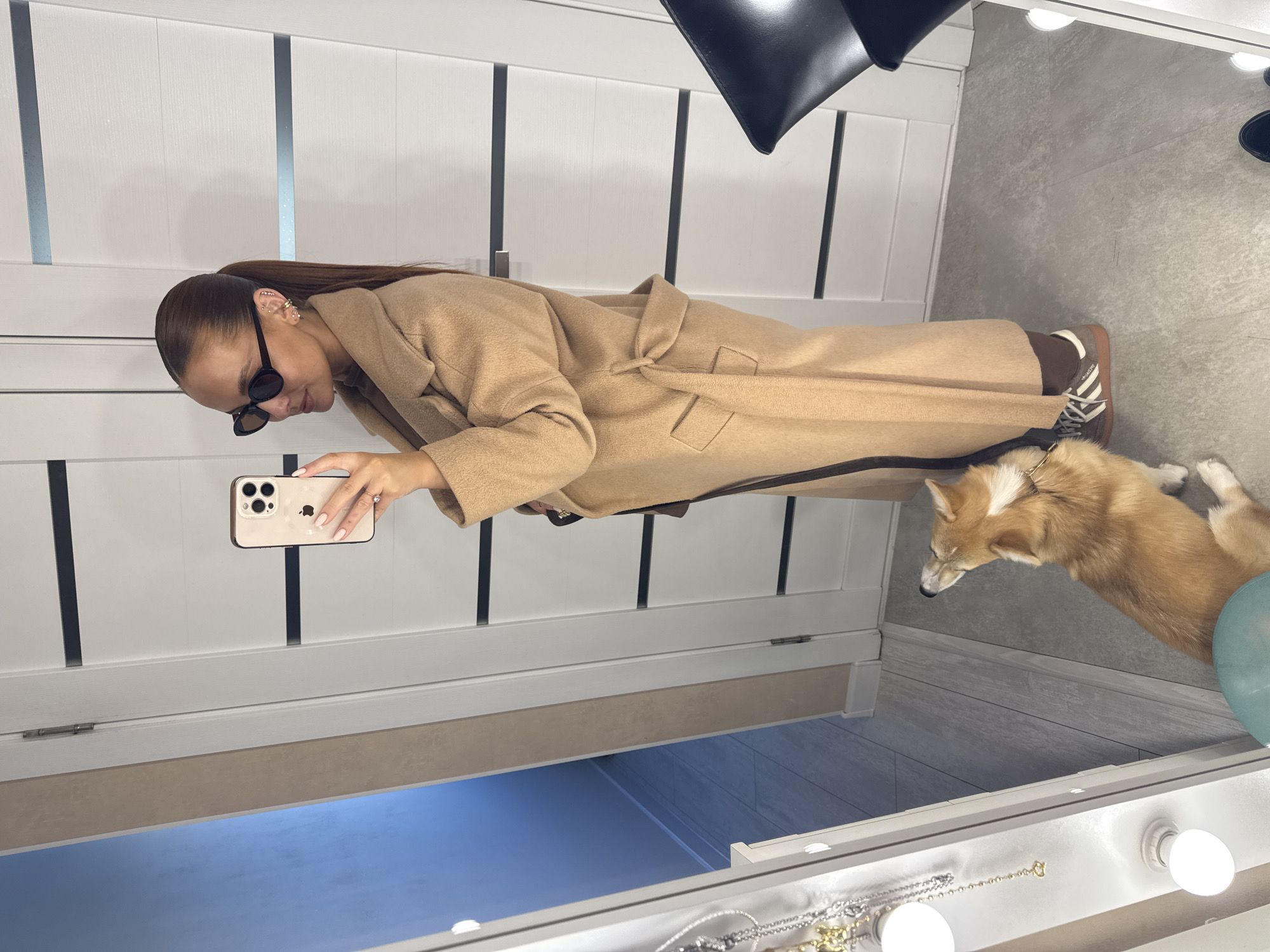 A woman with sunglasses and a light brown coat is taking a selfie with her phone. A dog lies on the floor next to her. The background is a wall with vertical white panels and a mirror with lights. The location may be in Chaikovsky, Russia, but this cannot be verified from the image alone.