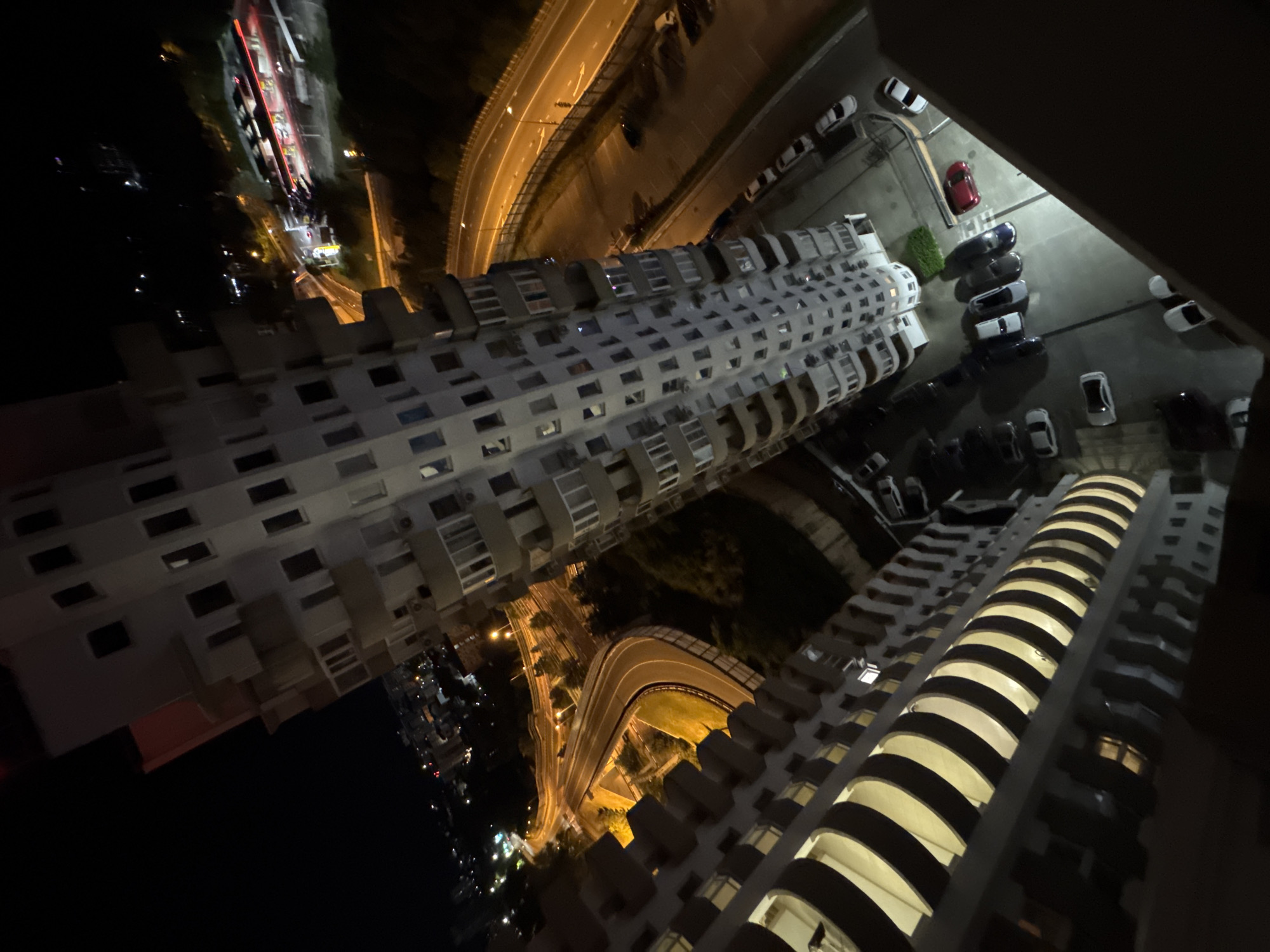 This is a nighttime, aerial view photograph taken from a high vantage point overlooking a complex urban environment in Sochi, Russia. The primary focus is on a tall, modern apartment building with numerous balconies, illuminated from within. Adjacent to this building is a multi-lane highway, with cars visible on the road and a red sports car parked in a lot below. There are other buildings and city lights in the background, indicating a densely populated area. The overall impression is one of a city at night, with residential and infrastructural elements clearly visible.