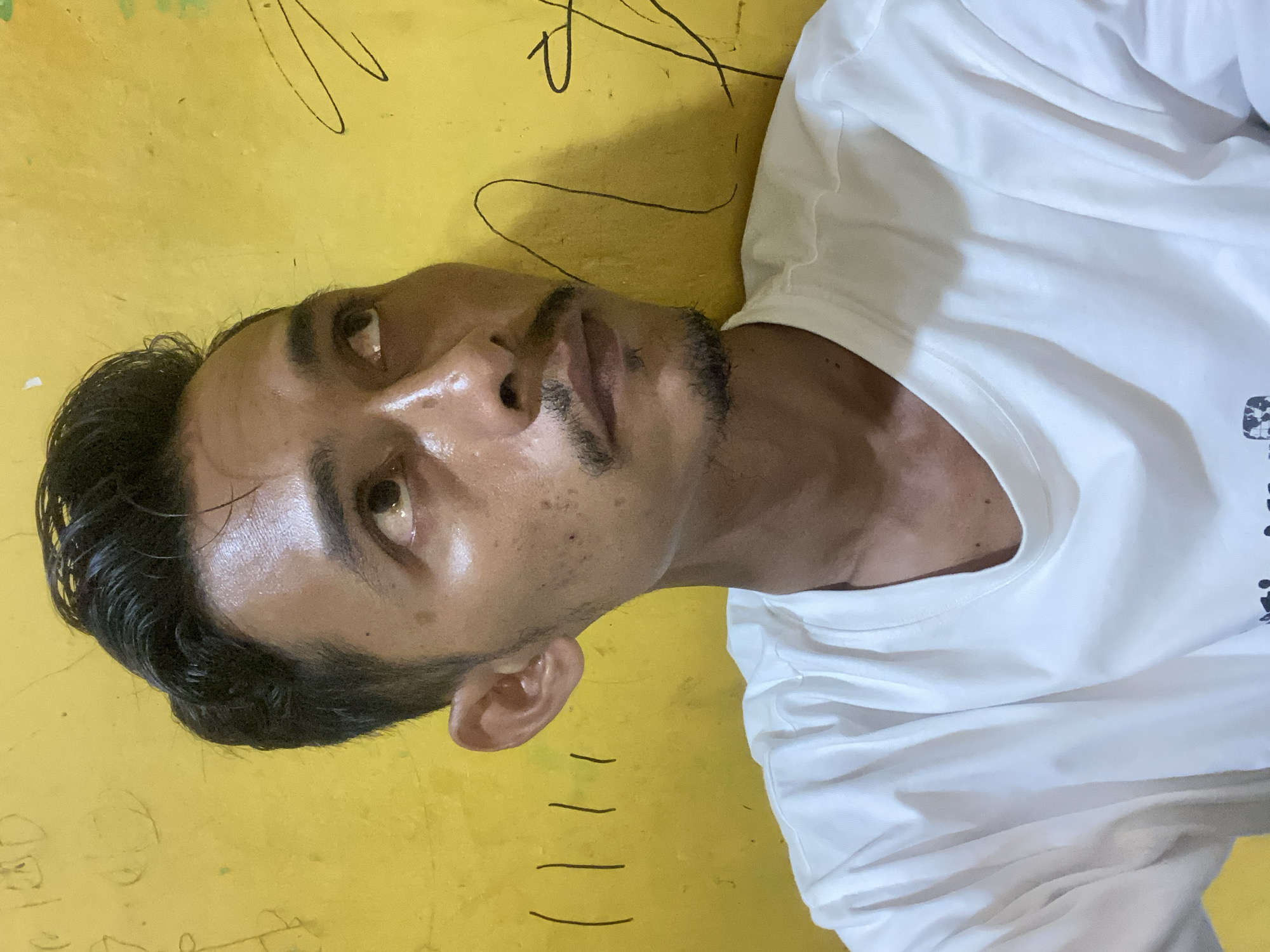 The image shows a close-up of a man lying down against a yellow wall in Tanjongkapai, Indonesia. The man is wearing a white t-shirt. He has dark hair and a trimmed beard. His skin appears to be sweaty or oily. The wall behind him has some markings or writing on it.