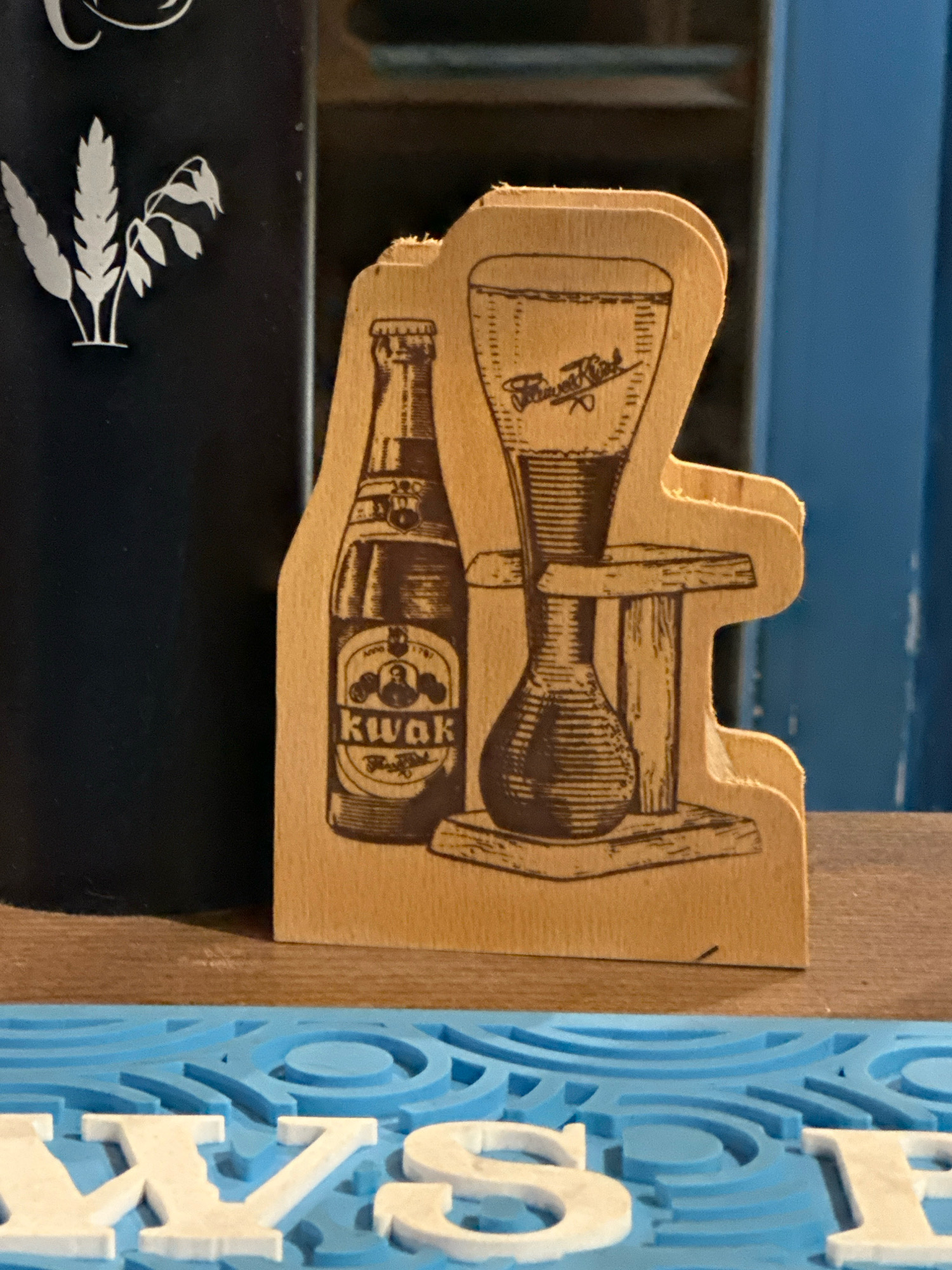 A wooden coaster with a laser-engraved image of a Kwak beer bottle and a Kwak glass on a wooden stand is positioned on a wooden bar top. The coaster is in the foreground, slightly to the right of the center. To the left of the coaster, partially visible, is a black object with a white stencil of wheat stalks and leaves. In the background, a blue vertical surface is visible, possibly a wall or cabinetry. The bar top appears to be made of wood with visible grain. A blue mat with embossed lettering is in the lower portion of the image, partially obscuring the bar top.