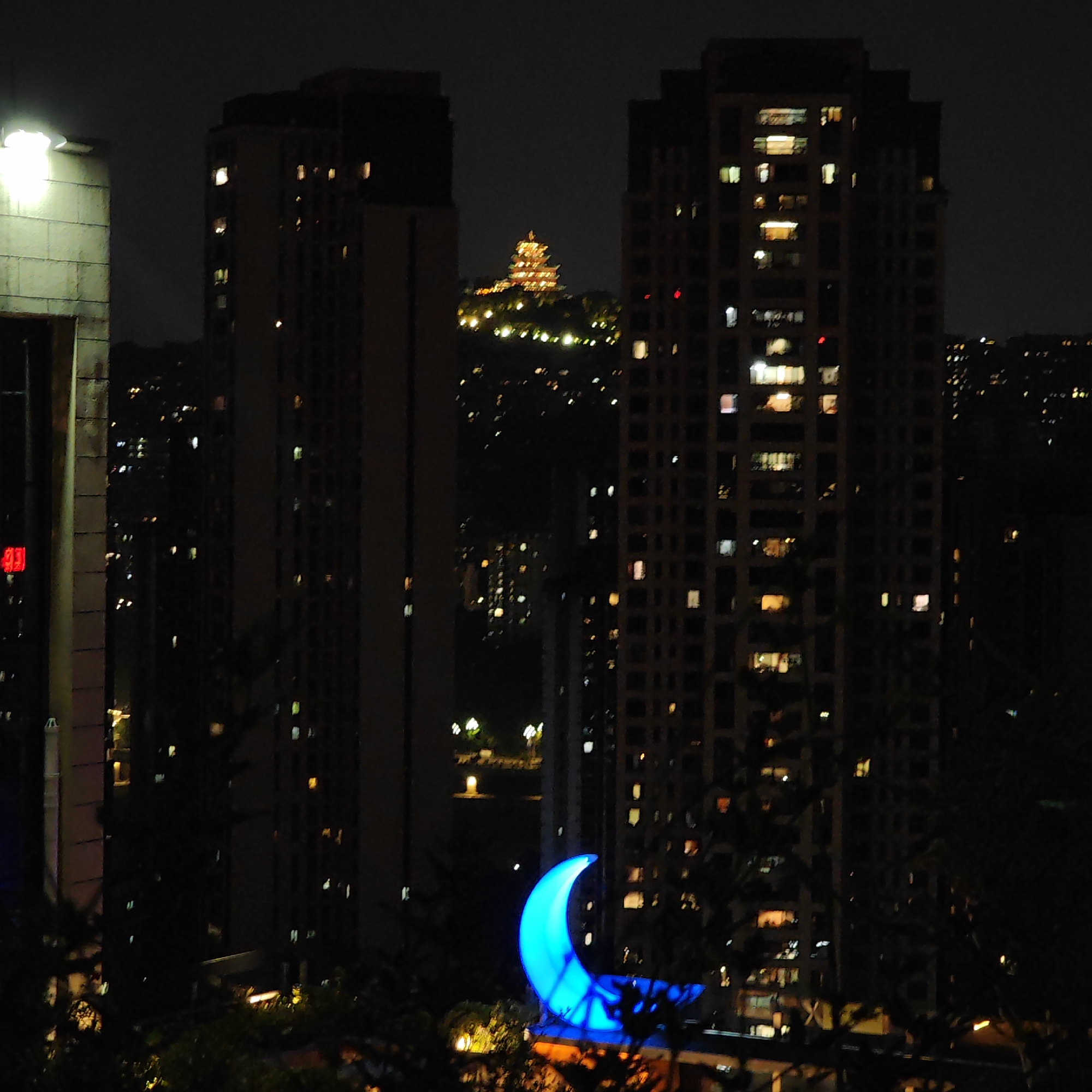 A nighttime view in Yuzhong District, China shows two high-rise buildings with illuminated windows. In the distance, a glowing structure sits atop a hill. A large, illuminated blue crescent moon is visible in the foreground. Other lights dot the cityscape. Tree branches and foliage frame the bottom of the view.