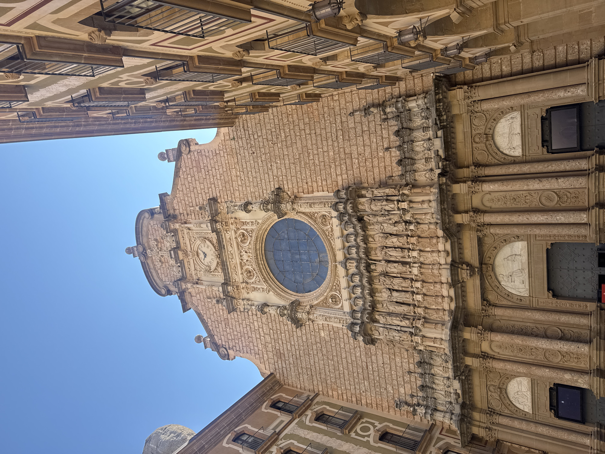 A low-angle outdoor shot captures the facade of Montserrat Abbey in Monistrol de Montserrat, Spain. The sandstone building is intricately detailed with numerous statues lining the upper facade, above a large, circular stained-glass window. A clock is visible above the window, set within a decorative stone frame. Flanking the abbey on the right is a multi-story building with balconies and decorative striping. On the left, part of another building is visible, with a large rock formation in the background. The sky is clear and blue.