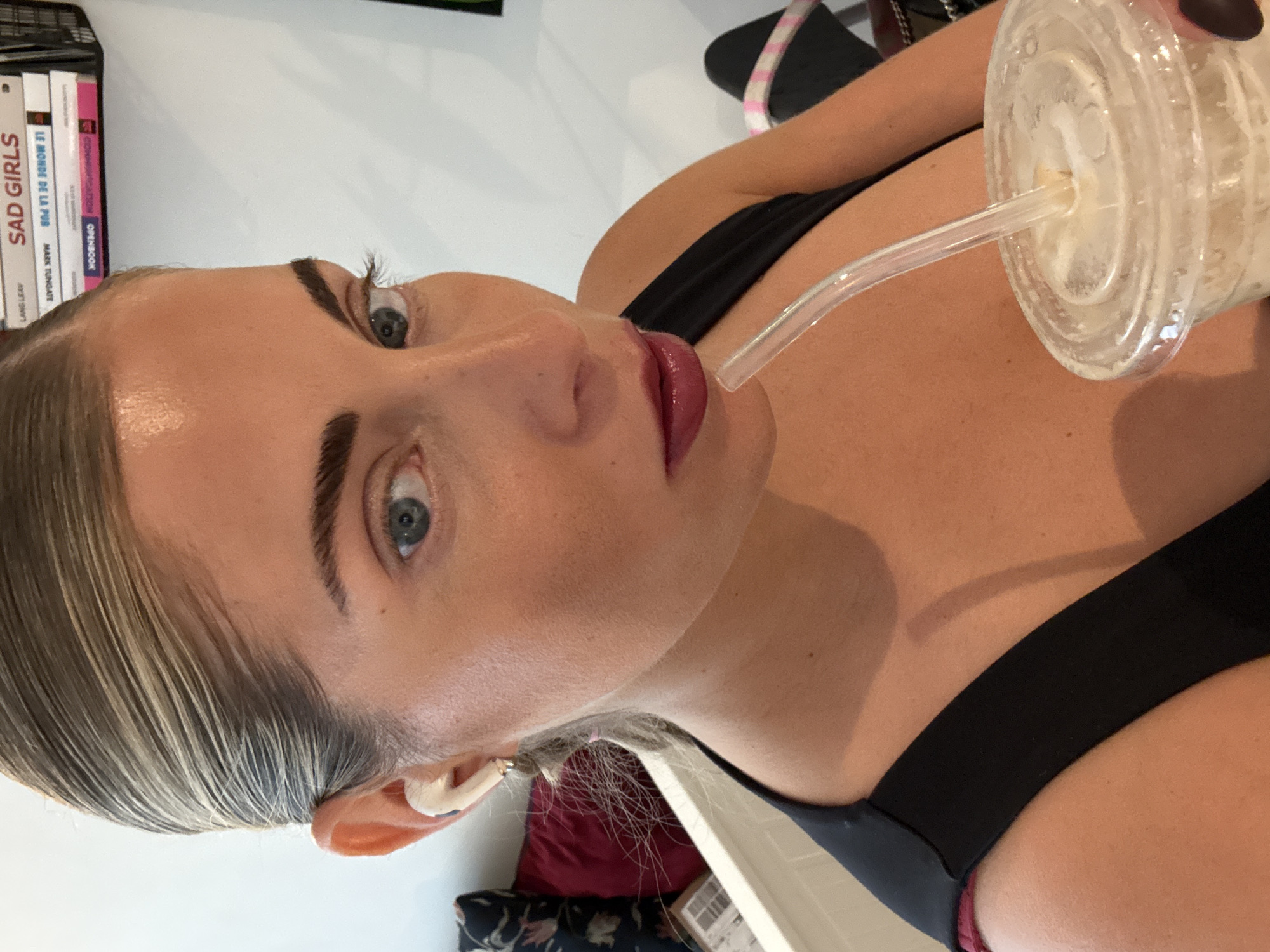 The image shows a fair-skinned woman with blue eyes, dark eyebrows, and blonde hair drinking from a clear plastic cup with a straw. She is wearing a black top. The background includes a stack of books with visible titles, including "Sad Girls" and "Le Monde de la Pub." The location is not discernible from the image.