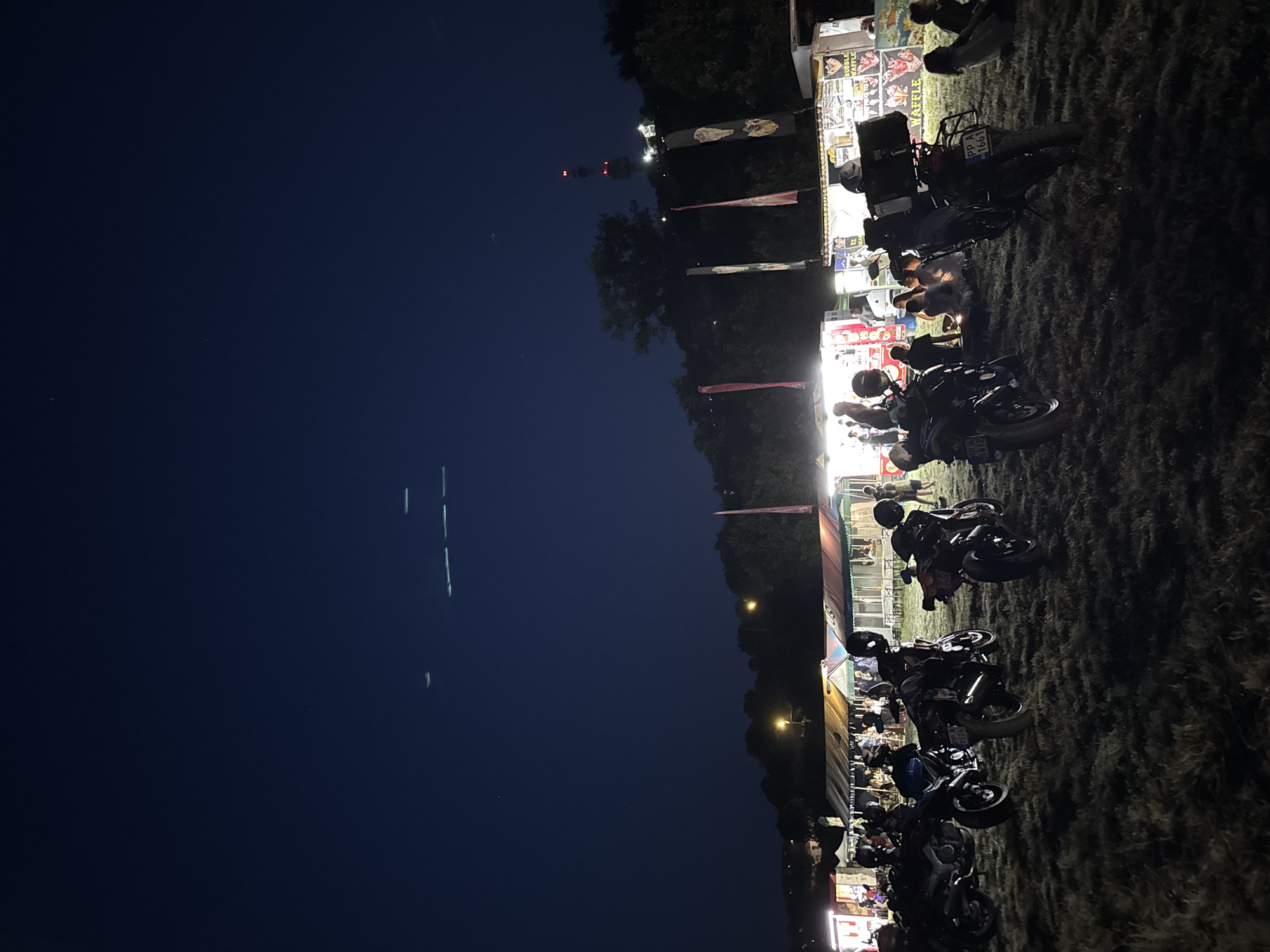 The image captures a nighttime outdoor event, likely a festival or gathering, situated on a grassy field. In the foreground, several motorcycles are parked, suggesting an event attended by motorcycle enthusiasts. Behind the motorcycles, there are rows of food stalls or vendor tents, illuminated by artificial lights, with people mingling and sitting on the grass, enjoying the atmosphere. In the background, a dimly lit tower with red and white lights is visible on a hill, identifiable as the Telecommunication Tower in Tutrakan, Bulgaria. The sky above is dark blue, with a few faint streaks of light, possibly from distant aircraft or atmospheric phenomena.