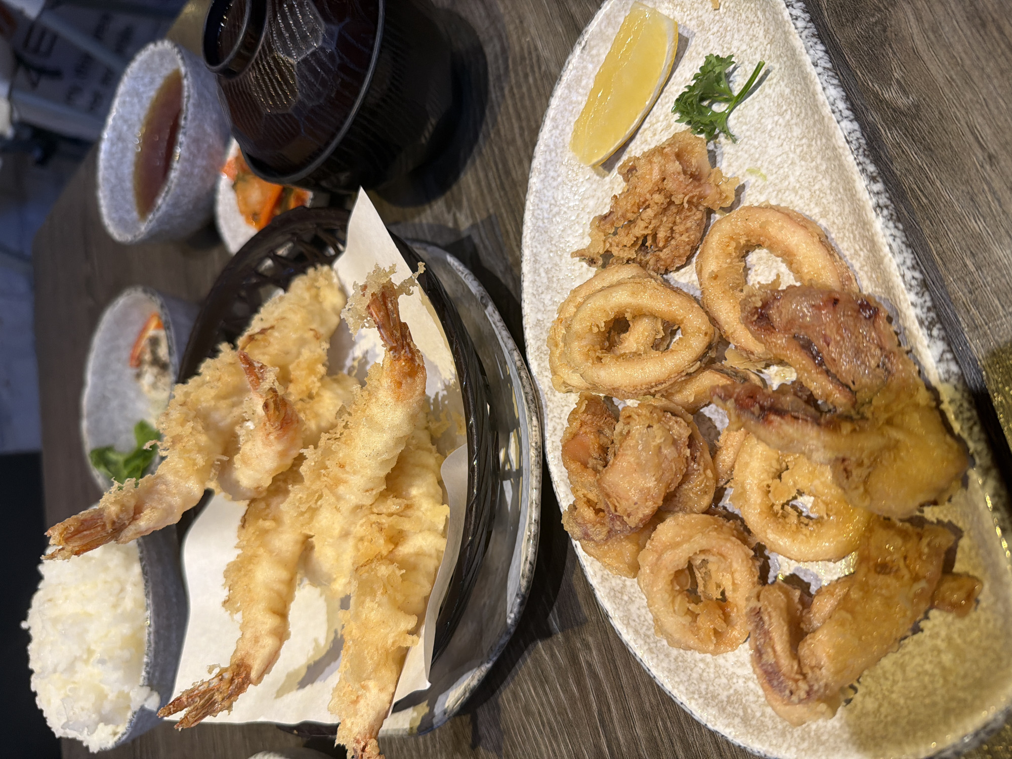 The image shows two plates of fried food on a wooden table. The plate on the left contains tempura shrimp and white rice. The plate on the right contains fried calamari and other fried seafood, accompanied by a wedge of lemon and a sprig of parsley. In the background, there is a small bowl of soup and a black bowl, as well as small items of food, partially visible. The location in the city of บ้านบางรักษ์, Thailand is not determined by the image.