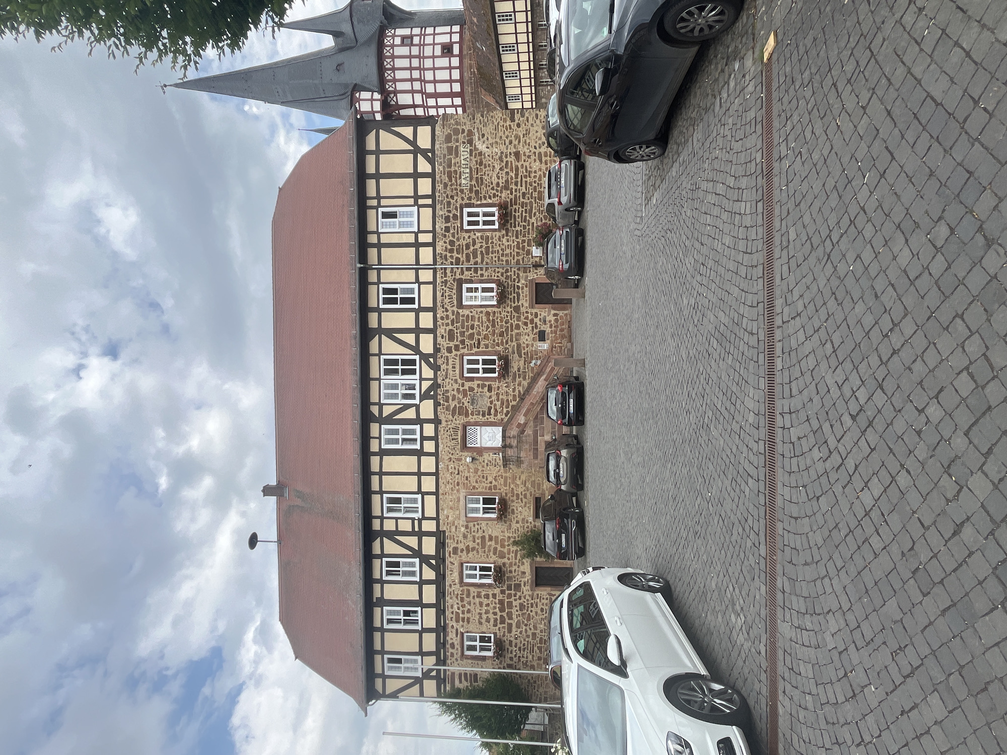 The image captures a street scene in Neustadt, Germany, featuring a historic building with timber framing and stone masonry. The building has a red-tiled roof and numerous windows with white frames. A sign on the building reads "Rathaus," indicating it is the town hall. Several cars are parked on the cobblestone street in front of the building. A white car is prominently positioned in the foreground, with its side and wheels visible. The sky is partly cloudy, suggesting it is daytime with mild weather. The scene appears to be a typical street view in a German town, with traditional architecture and functional parking. There are no people visible in the image, and the overall impression is one of a quiet, everyday moment.