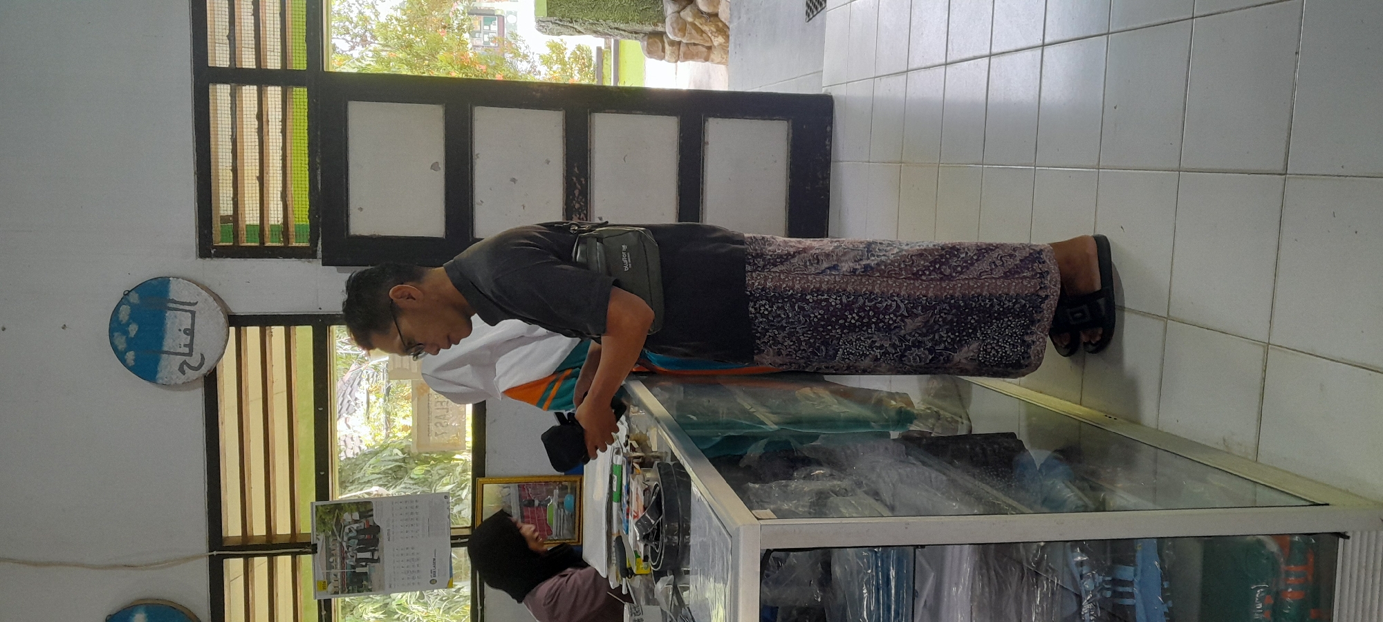 In Tahunan, Indonesia, a man wearing a dark t-shirt, a sarong, and glasses stands at a counter, holding a black object. He has a small, dark green bag slung across his chest. Behind him, a woman wearing a black hijab is partially visible, looking down and smiling. Another person, wearing a white hijab and an orange and blue garment, stands between them. The counter is filled with various items, including what appear to be pens, a roll of tape, and a black belt. In the background, a calendar with a picture of people in uniform is displayed on the wall. Above the window, a round decoration with Arabic script and blue clouds is mounted. The room has tiled floors and a wooden door with multiple glass panels.