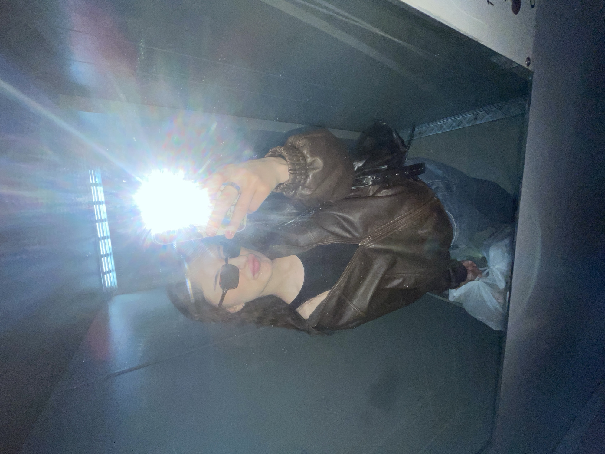 A woman wearing a brown leather jacket, black top, and sunglasses is taking a selfie with a bright light source, possibly a phone, inside what appears to be a container or confined space with metallic walls and a grayish floor. A plastic bag and other objects are visible in the background. There is no information given in this image that indicates the location is Moscow, Russia.