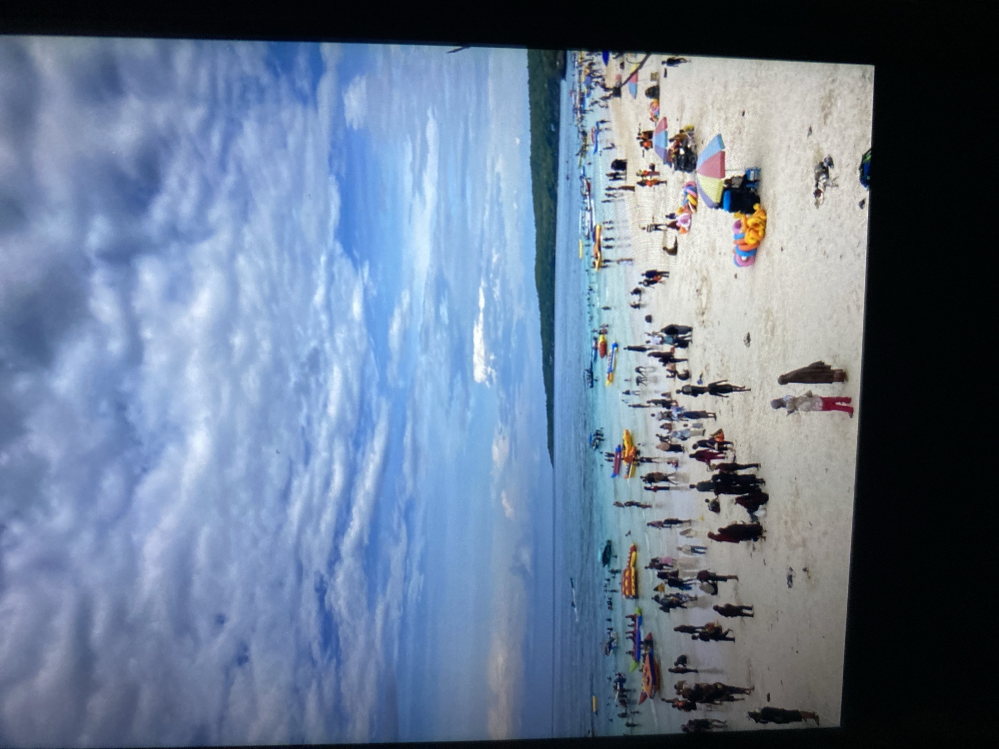 The image depicts a crowded beach scene on a cloudy day, likely in the city of Ta', Indonesia. The beach is populated with numerous individuals engaged in various activities. In the foreground, people are walking on the sand and gathered in small groups. Some are wearing casual beach attire, while others are in modest clothing, including headscarves.
Further into the scene, the shallow water is filled with people swimming and wading. Various inflatable watercraft, such as banana boats and tubes, are visible both in the water and on the beach, suggesting recreational activities. Boats are also present in the sea, some of which appear to be small passenger vessels.
The environment consists of a sandy beach bordering a clear blue sea, with a lush, green, tree-covered landmass in the background. The sky is predominantly covered with white and grey clouds, with patches of blue sky peeking through. Beach umbrellas and chairs are scattered on the sand, indicating a typical tourist destination. The overall impression is one of a busy, tropical beach during daylight hours.