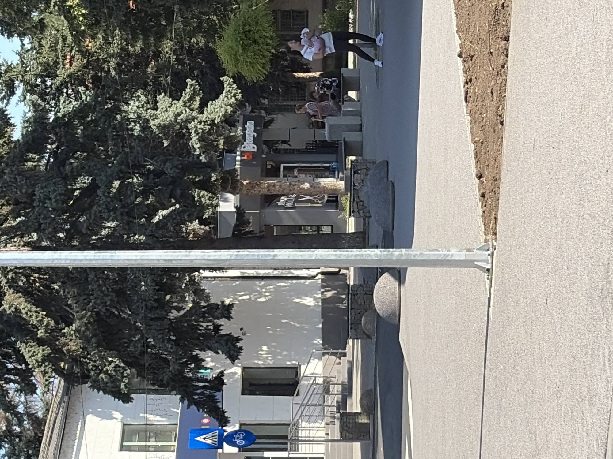The image depicts an outdoor urban scene, likely in Criuleni, Moldova, on a sunny day. In the foreground, a paved area with a concrete curb and a patch of disturbed earth is visible. A light-colored, smooth, rounded bollard is present on the pavement. A thin, vertical metal pole, possibly a lamppost, stands prominently in the mid-ground, casting a shadow.
Behind the pole, a building with a white facade and a green-framed window is partially obscured by a large evergreen tree with dense foliage. A blue traffic sign displaying a pedestrian symbol and a bicycle symbol is affixed to the building. Further back, another building with dark storefront windows and signage, one reading "Play.auto," is visible.
Several people are present. On the right side, a woman in a white t-shirt and black leggings is holding a baby wrapped in a pink blanket. She is walking away from the camera. In the mid-ground, two people are seated on a stone bench in front of the storefront. Another person is standing near them.
The environment includes more evergreen trees, and the ground is paved with a light-colored material, possibly asphalt or concrete. The overall impression is a public space, possibly a pedestrian zone or a plaza, adjacent to commercial buildings and residential structures.