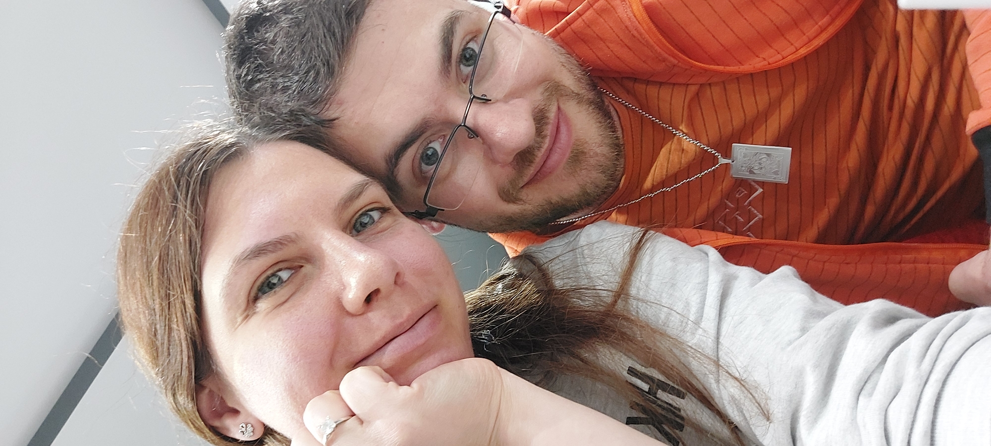 A selfie shows a woman and a man. The woman has brown hair and is wearing a grey sweater and a ring. She has her hand under her chin. The man, who is behind her, has short dark hair, glasses, and a beard. He is wearing an orange shirt with a pendant. No location information is inferable from this image.
