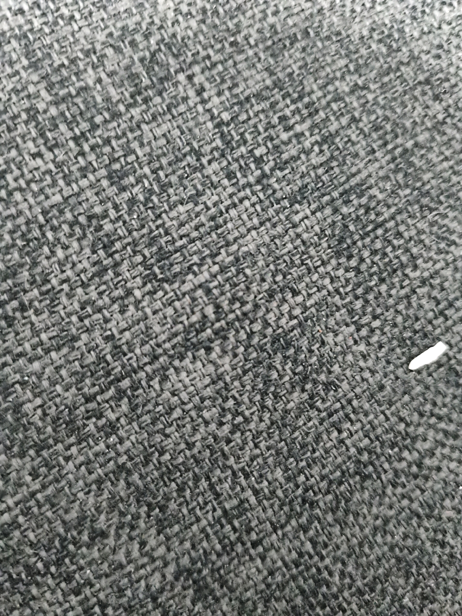 A close-up, high-angle shot shows the textured surface of a dark gray woven fabric. The weave consists of tightly interlaced threads, creating a pattern of small, rectangular openings. The fabric appears to be a single, continuous surface. There are no discernable objects or people within the frame. The image is taken in Sukasejati, Indonesia.