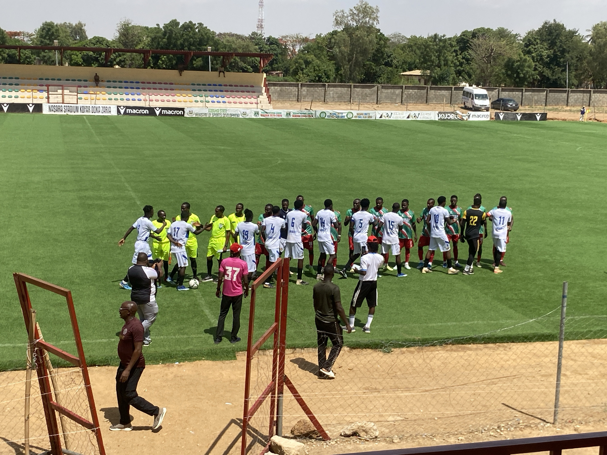 The image depicts a soccer match taking place at the Nasara Stadium Kofar Doka Zaria in Tudun Wada, Nigeria. Two soccer teams in their kits (one in white/blue and the other in green/red) stand on a vibrant green field. Several individuals dressed in contrasting clothing are also present including some in yellow and a man in a pink shirt. Several officials and bystanders stand near the teams. The soccer field is surrounded by a fence. Behind the fence is a partially visible stadium with colorful seats and stadium advertising. A low wall borders the stadium, and vehicles are parked beyond it. A grassy field and trees surround the area. In the foreground is a red frame and fence.