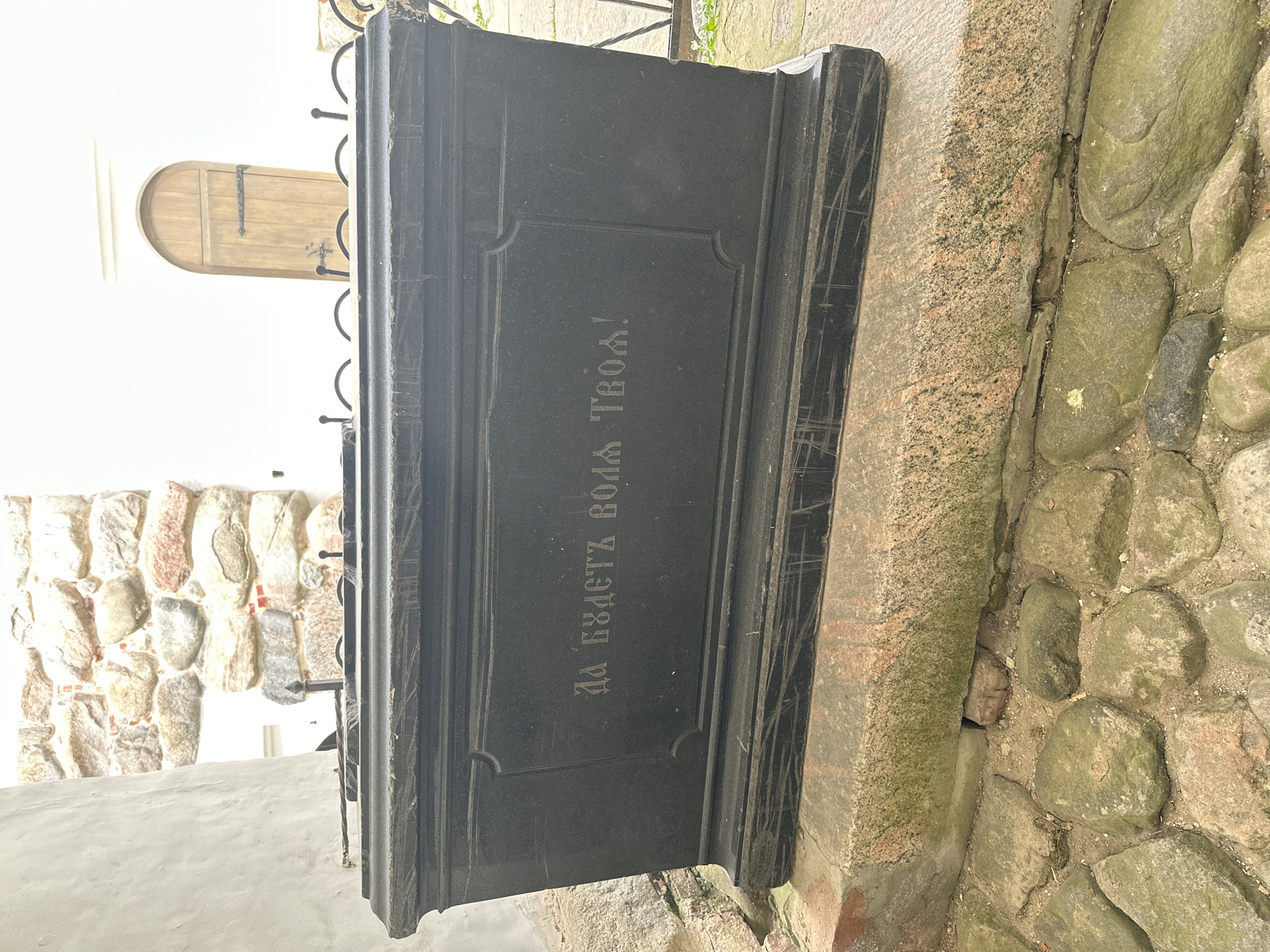 The image shows a dark granite sarcophagus situated on a stone foundation, with cobblestones in the foreground. The sarcophagus has an inscription in Cyrillic letters on its front face, reading "Да будет воля Твоя!" which translates to "Thy will be done!". In the background, a stone wall with large, rough-hewn stones is visible, along with a white building and a wooden arched door. The sarcophagus is located at the Solovetskiy Monastery in Russia. The lighting suggests it is daytime, possibly overcast, as there are no strong shadows. No people are present in the frame.