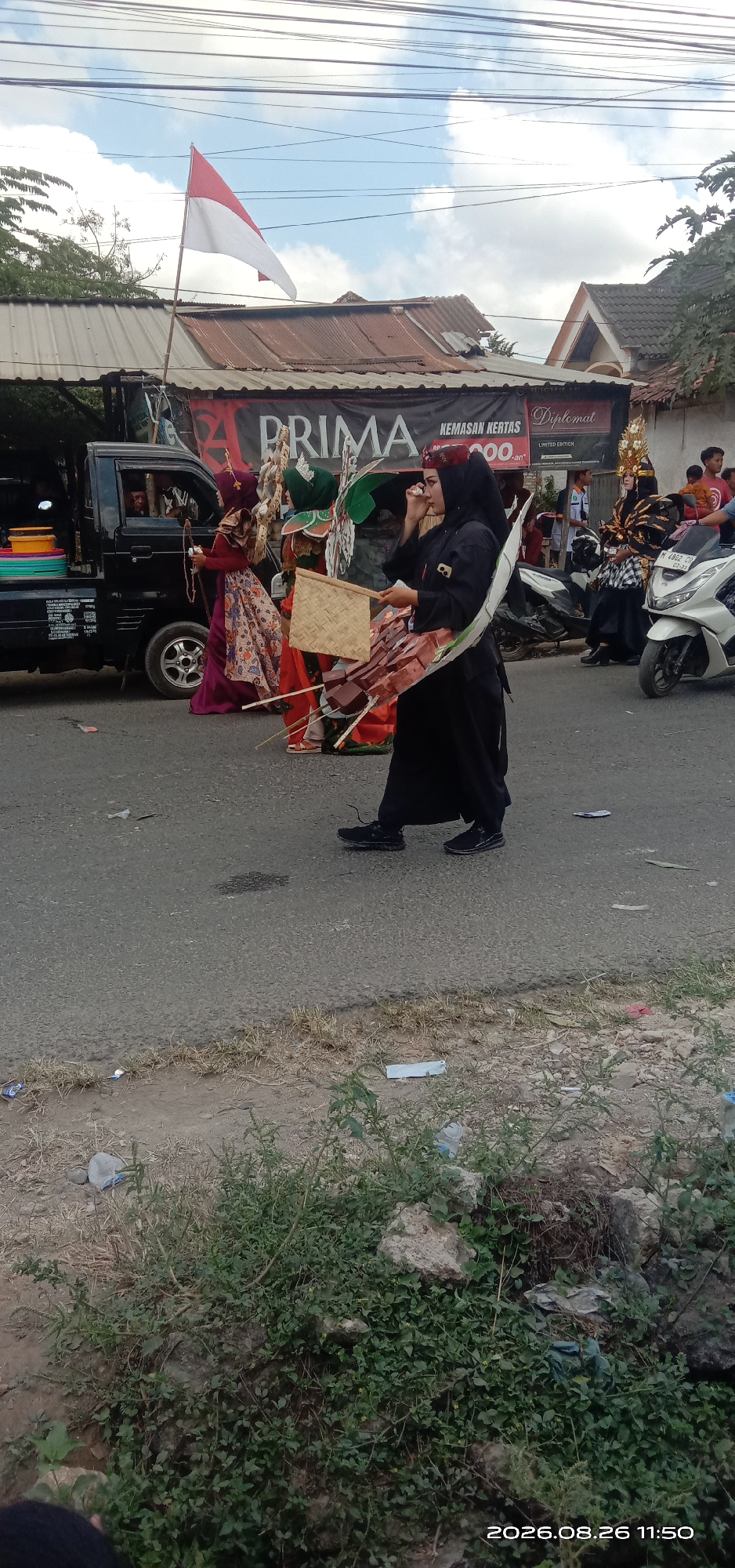 In the city of Pakong, Indonesia, a procession is underway. A woman in a black abaya and headscarf walks on the right side of the road, holding a fan-like object. Another person in traditional Indonesian attire, adorned with a gold headdress and colorful fabric, stands behind her, observing. To the left, a black pickup truck is parked, its side visible, with people inside. A red and white Indonesian flag flutters from a pole mounted on the truck. The background shows buildings with corrugated roofs and a banner with "PRIMA" written on it, along with other text and a logo. Several motorcycles are parked on the right side of the road. The scene appears to be a cultural event or celebration taking place on a paved road with grassy areas and some debris on the sides.