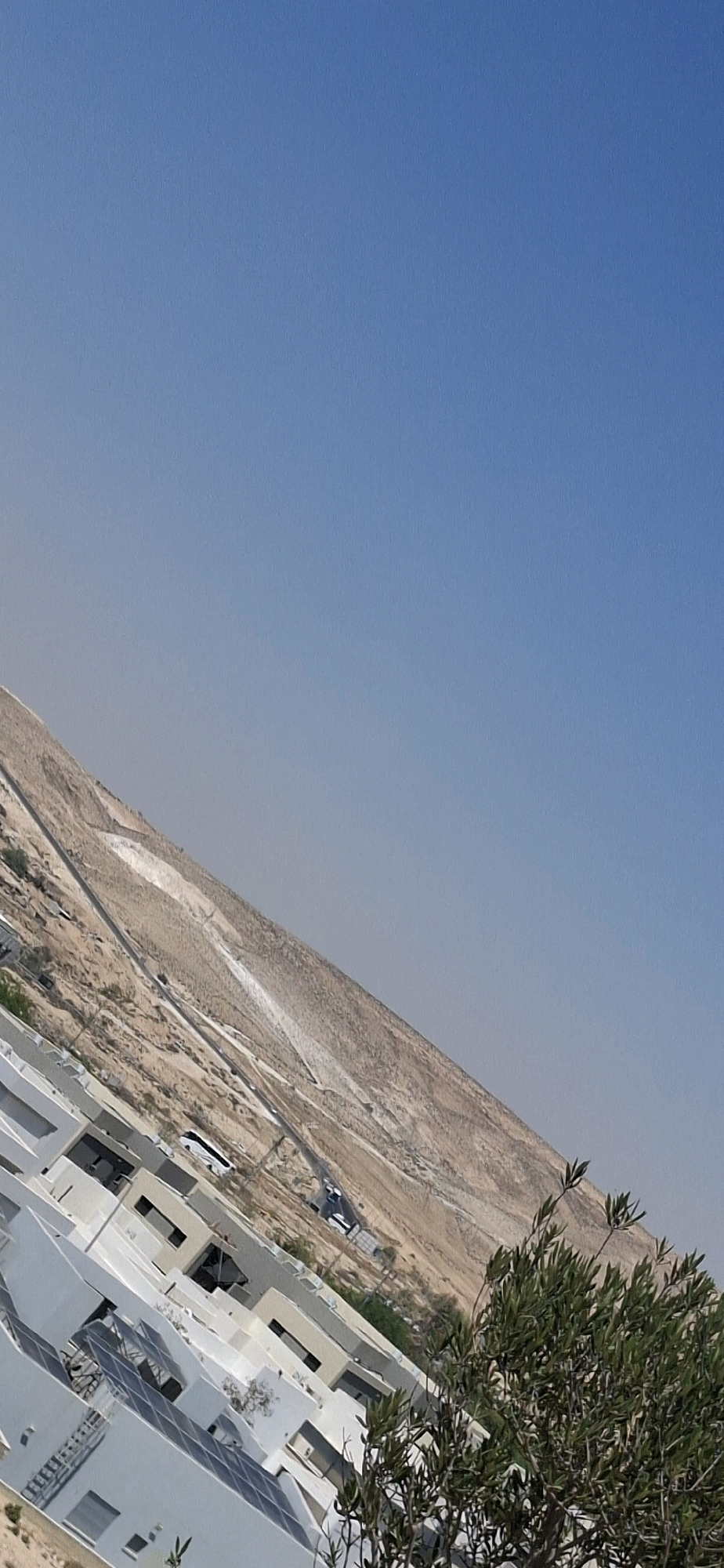 The image is a tilted, medium-distance shot of a residential area in Yerocham, Israel, with a prominent desert hillside in the background. In the foreground, modern, white, multi-story buildings with flat roofs and numerous windows are visible. Some rooftops are equipped with solar panels. A sparse, green shrubbery is present on the right side of the frame. In the mid-ground, a road winds its way up a light brown, barren hillside. Several white vehicles, including what appears to be a bus, are traveling on this road. The sky is a clear, light blue. The overall impression is one of a modern settlement at the edge of a desert landscape.