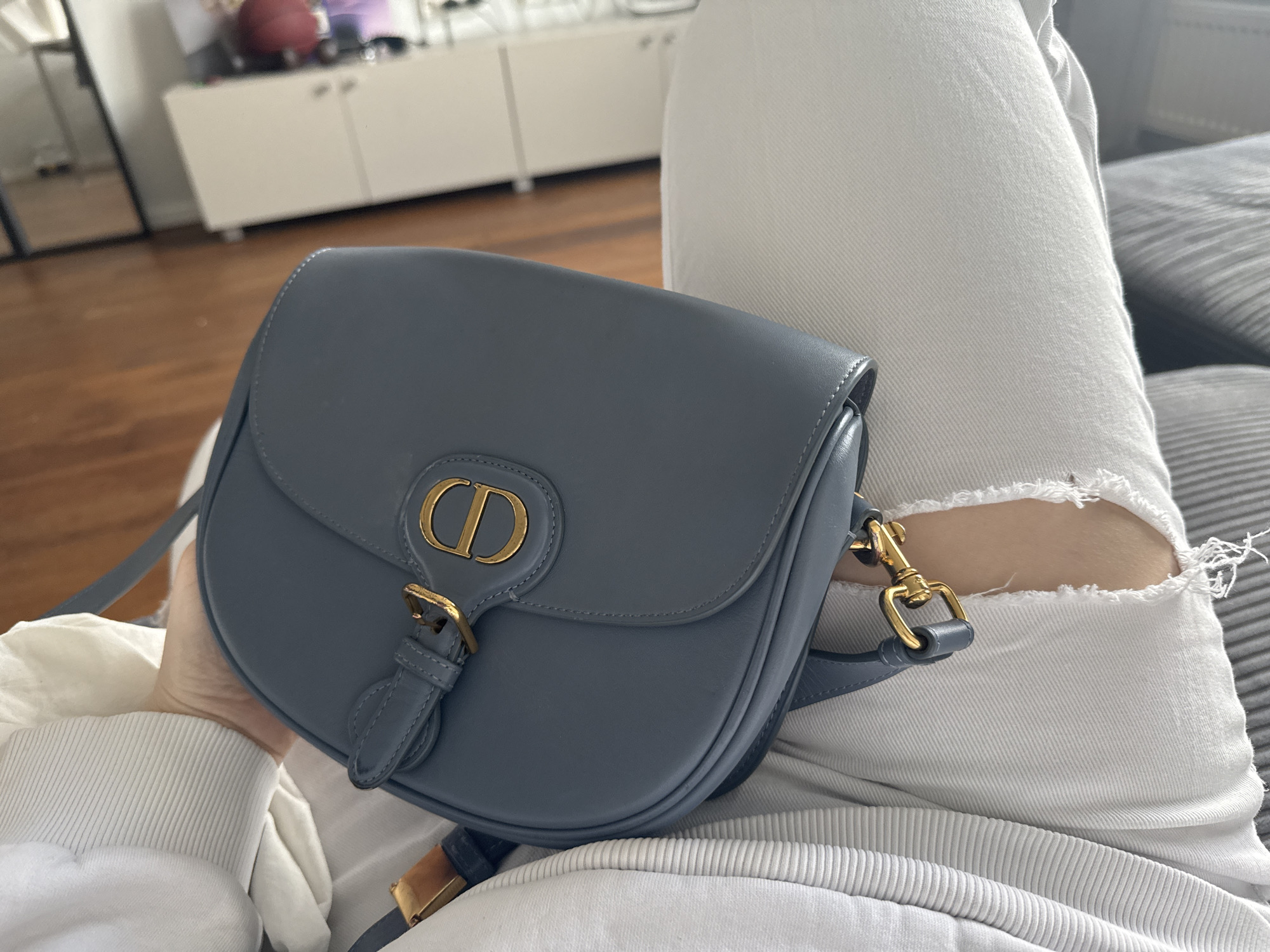 A person wearing white ripped jeans and a white long-sleeved shirt is holding a blue Dior bag on their lap. The background shows a light brown wooden floor and a white cabinet with a few items on top. A grey sofa is partially visible on the right. The location in the city of Khimki, Russia is not discernible from this image.