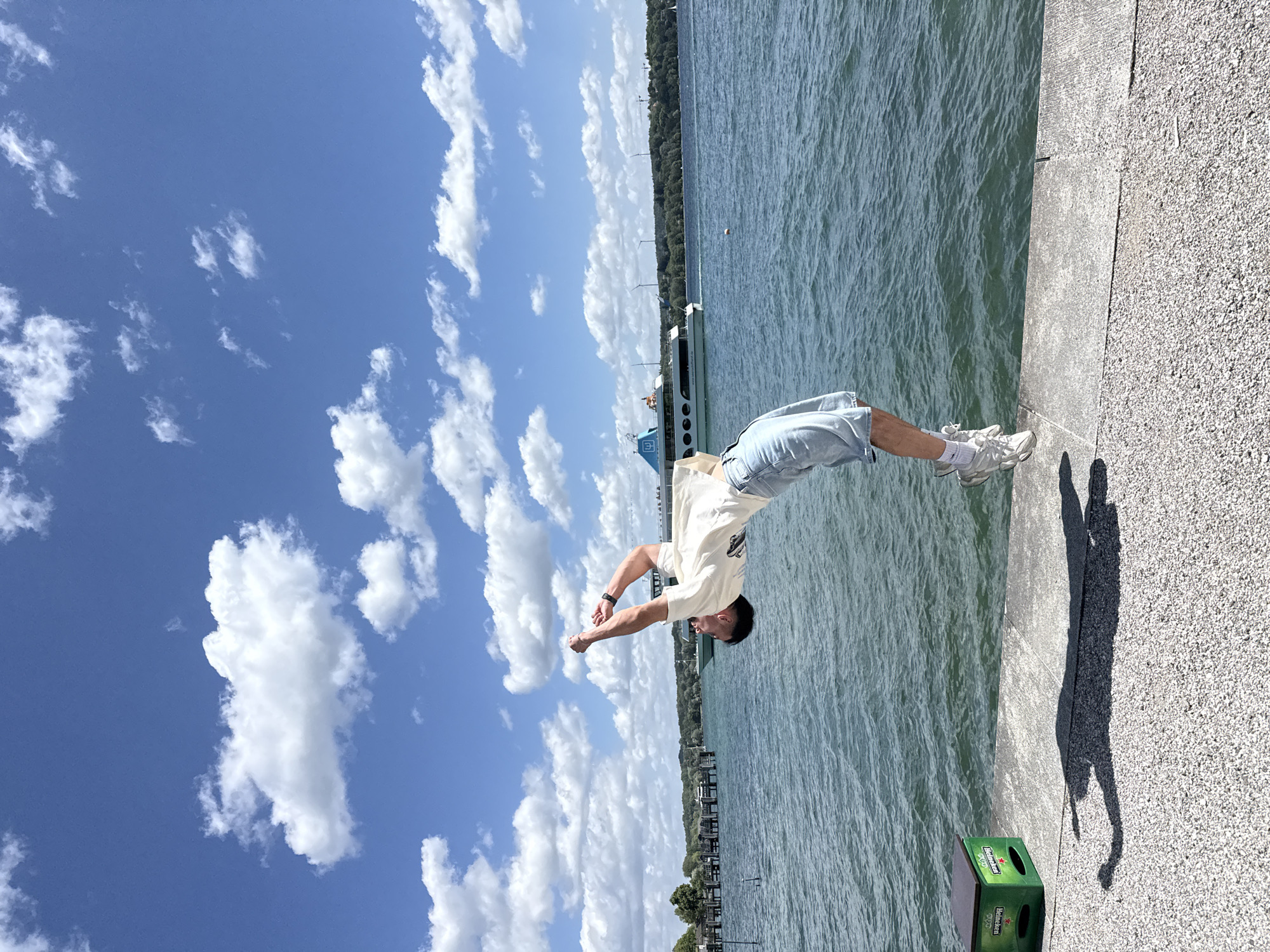 A young man is captured mid-flip in the air against a backdrop of a blue sky with scattered white clouds, a body of water, and a distant shoreline. The man is wearing a cream-colored t-shirt and light blue denim shorts, with white sneakers. He is arching backward, with his arms raised and hands clasped. The scene appears to be on a paved walkway next to a large body of water, possibly a lake. In the background, a white and blue passenger boat is visible on the water, and the shoreline is lined with trees and some structures. A green crate with "Heineken" branding is on the left side of the walkway. The overall impression is of a dynamic, outdoor activity taking place on a sunny day. This location is the Starnberger See in Starnberg, Germany.