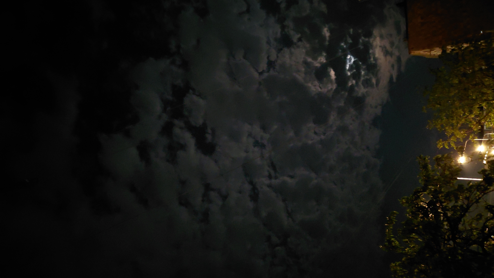 The image depicts a nighttime scene with a dark, cloudy sky. In the lower portion of the frame, trees are visible, partially illuminated by streetlights. A portion of a building with a reddish-brown roof is present on the right side. The moon is faintly visible through a break in the clouds in the upper right quadrant. The scene is urban, likely in Abovyan, Armenia, given the context. There are no people or specific actions depicted. The environment is characterized by darkness, clouds, and artificial lighting from street lamps.