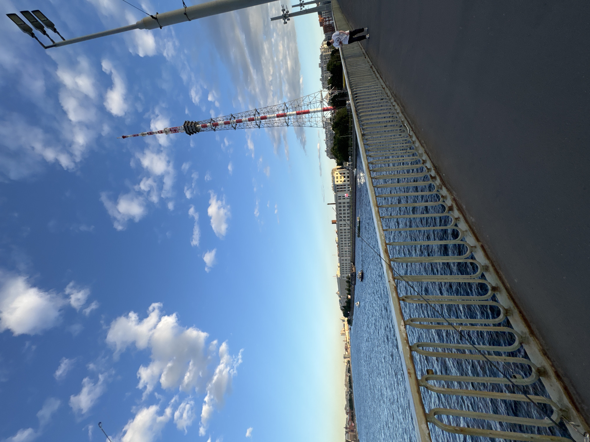 The image depicts a wide, dark asphalt pedestrian walkway on a bridge, bordered by a weathered metal railing on the left. The railing has a light beige top rail with noticeable rust streaks and a decorative pattern of vertically oriented, looped metal bars. Beyond the railing, a wide, dark blue river fills the frame, with ripples indicating a light breeze. Several boats are visible on the water. Across the river, the cityscape of Saint Petersburg, Russia, stretches along the horizon. Notable buildings include a long, white, multi-story structure with numerous windows and a red-tiled roof. Further in the distance, more urban architecture and a suspension bridge can be seen. Towering over the city, a tall, lattice radio tower with red and white stripes is prominently featured against a bright blue sky dotted with fluffy white clouds. A single street light with three fixtures stands on the right side of the bridge. A lone individual, a person wearing a white t-shirt and dark pants, is walking away from the camera on the right side of the walkway. The lighting suggests it is late afternoon or early evening, with the sun casting a gentle glow.