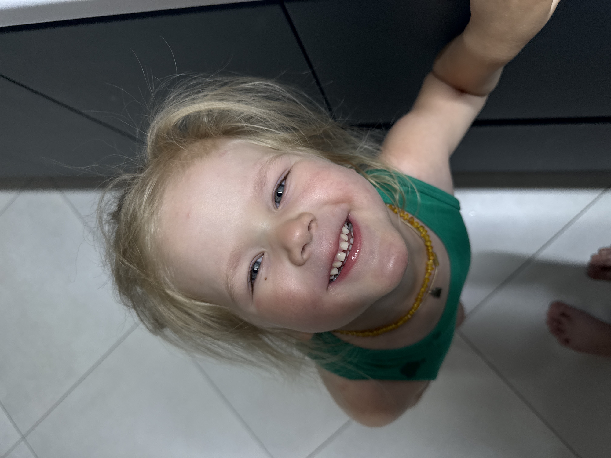 A young child with fair skin, blonde hair, and a green shirt is smiling directly at the camera. They are wearing a yellow beaded necklace. The child is reaching towards a dark, possibly black, surface above them. The floor beneath them is tiled in a light color with a grid pattern. A pair of bare feet are visible in the lower right corner of the frame. The photograph appears to be taken from a high angle, looking down at the child. The location is described as the city of Ферма, Russia, although there is no evidence of this in the image.